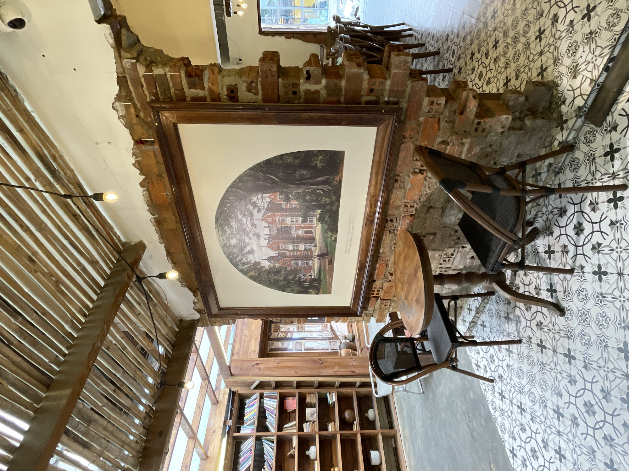 The image depicts an interior scene of a cafe with a rustic and exposed brick aesthetic, likely located in Phường Xuân Hương, Đà Lạt, Vietnam. The foreground features a small, round wooden table with two dark-colored chairs. The table is set on a patterned tile floor with a repeating geometric design in shades of gray and white. Behind the table, a large, framed artwork displaying a historical building surrounded by trees is mounted on a partially exposed brick wall. To the left of the artwork and table, a tall wooden bookshelf is filled with various books and decorative items. The ceiling features exposed wooden beams and bamboo slats, with two hanging light bulbs visible. In the background, a row of dark wooden chairs and tables are arranged, suggesting a larger seating area. The overall ambiance is casual and inviting, with elements of natural materials and vintage decor. There are no people visible in the scene.