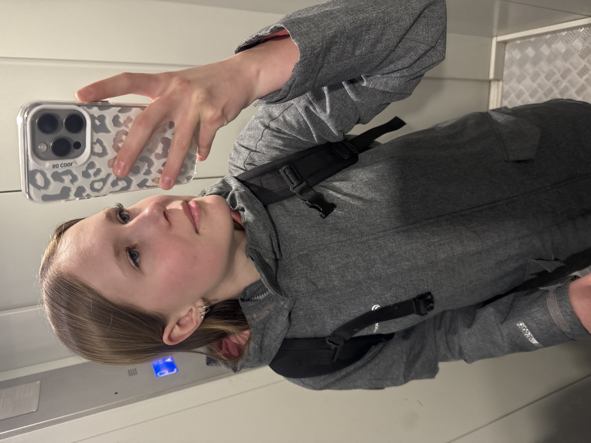 A fair-skinned young person with light brown hair takes a selfie in what appears to be an elevator. They wear a gray jacket and carry a black bag across their body. The phone case has a leopard print design. The elevator interior consists of white walls and a metal panel with a textured pattern. The location in Chaikovsky, Russia, cannot be determined from the image.