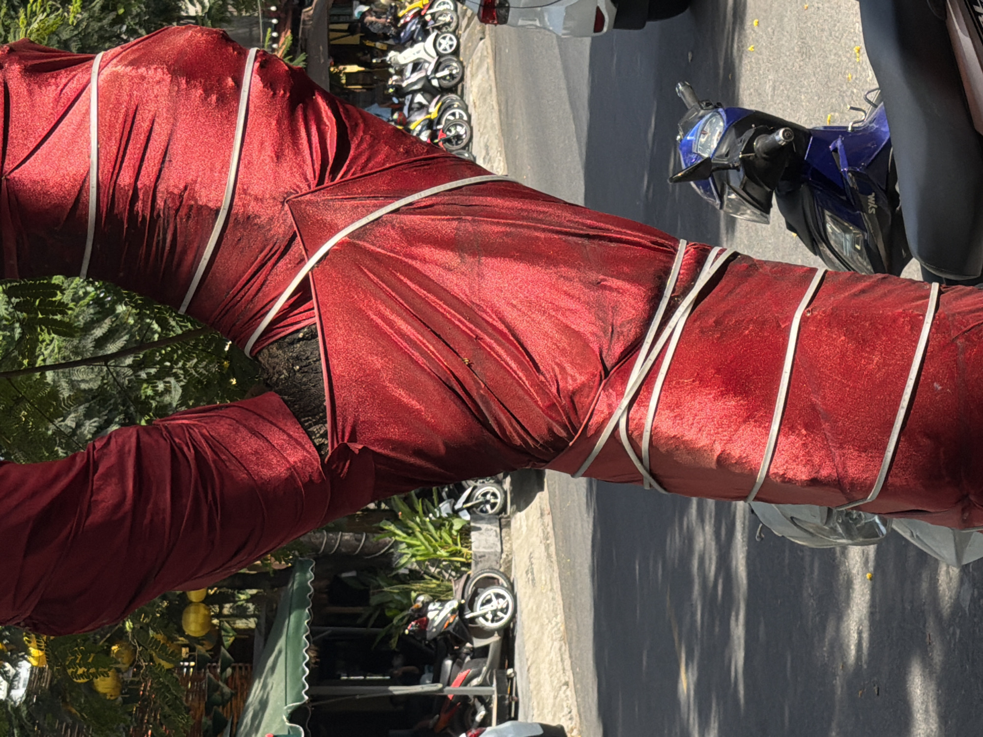 A tree wrapped in red fabric and secured with white tape stands near a street in Đà Nẵng, Vietnam. Several motorcycles are parked alongside the road and under the tree.
