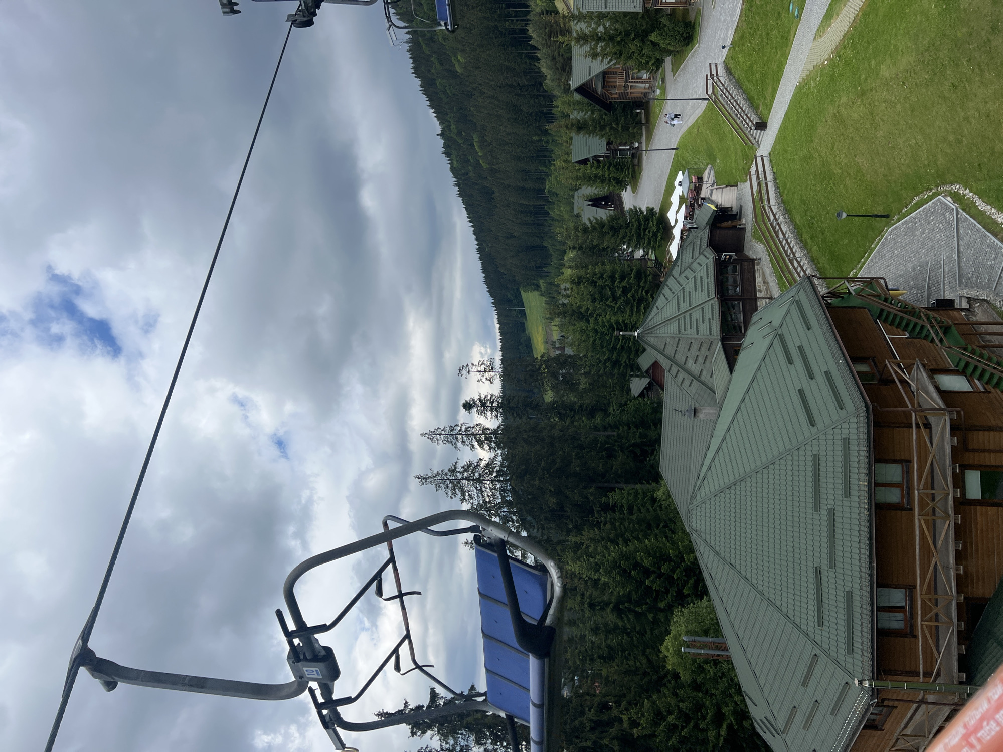 A ski lift chair with blue seats is visible in the foreground, suspended above a forested mountain resort. The resort features several wooden chalets with green roofs and is nestled among dense evergreen trees. A paved path winds through the resort, with a few people walking on it. The sky is cloudy, with patches of blue visible. The scene suggests a scenic, mountainous location, possibly a ski resort during the off-season or summer months.