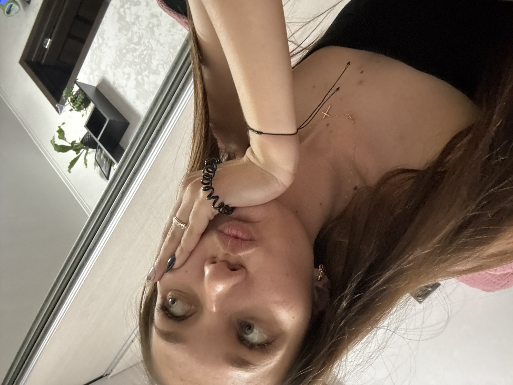 The media file is a close-up, vertically oriented selfie of a young woman with long, brown hair. She is wearing a black top and a gold cross necklace. Her right hand is touching her face, and she has a ring on her left index finger and a black bracelet on her right wrist. Her left eye is looking slightly to the left, and her lips are pursed. The background appears to be a mirror reflecting a wall with a patterned texture, a dark shelf with plants, and a doorway. The lighting suggests it is daytime. The overall emotion conveyed is playful or pouty. The scene is indoors, likely a home environment. No visible text is present. The provided location context is Novokuznetsk, Russia.