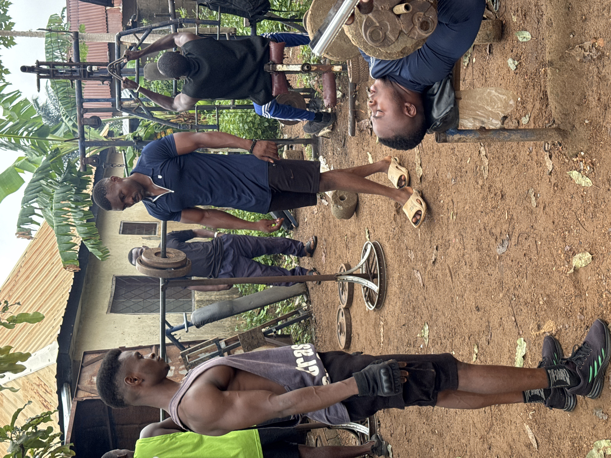 In Akwanga, Nigeria, an outdoor gym scene unfolds. Four young Black men are engaged in weight training. The foreground shows one man lying on a bench, lifting weights, with another man standing behind him, appearing to spot him. To the left, a man in a gray tank top and black shorts is standing, also wearing black gloves. In the background, another man is seen working out on a weight machine. The gym is set against a backdrop of rustic buildings and lush green foliage, suggesting a semi-urban or rural setting. The ground is covered in reddish-brown soil, scattered with leaves. The overall atmosphere depicts a communal workout session in a simple, open-air fitness environment.