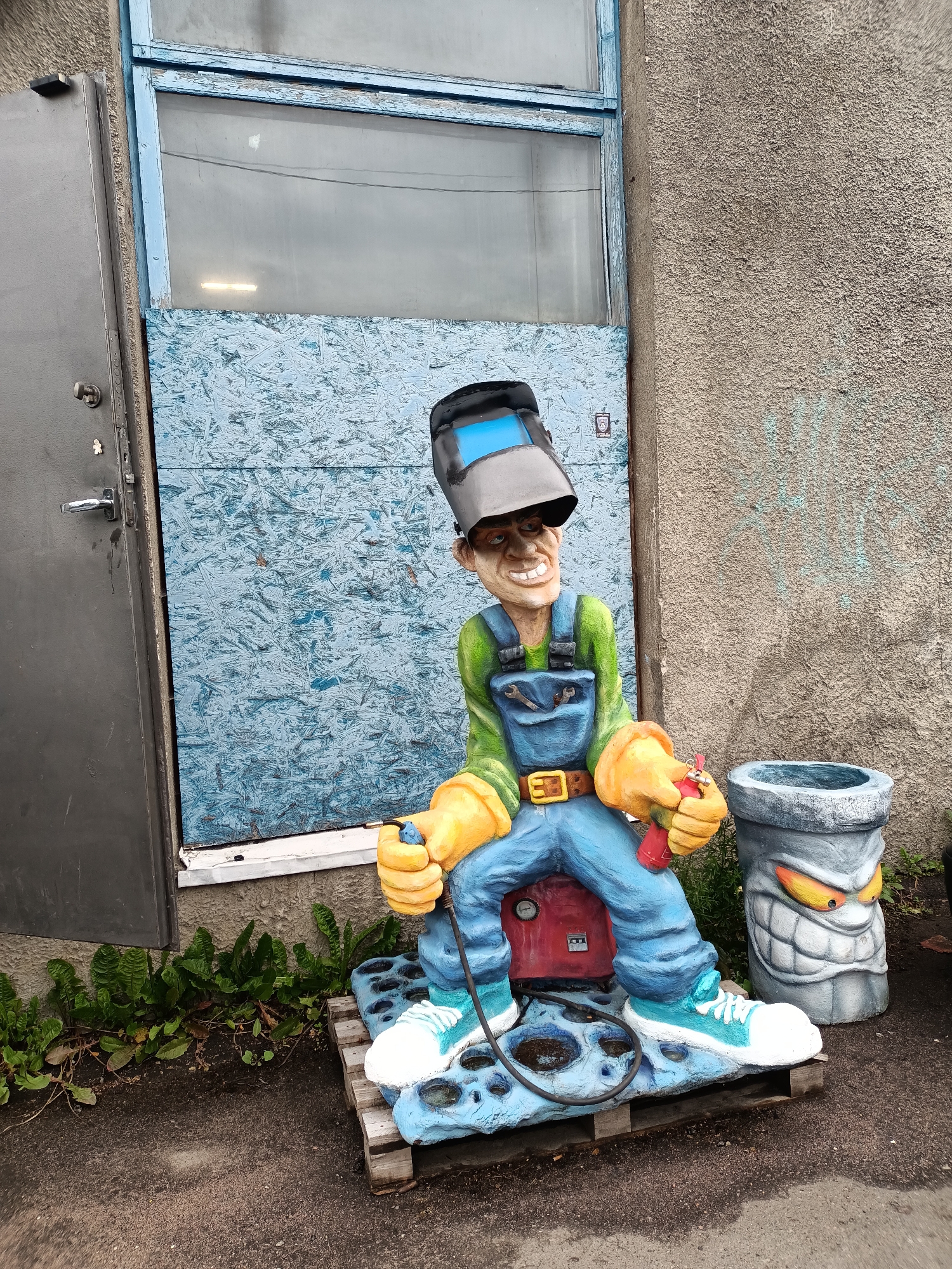 A life-sized, brightly colored statue of a caricatured welder sits outside a building in Tallinn, Estonia. The welder, with a welding mask tilted back on its head, green shirt, blue overalls, and yellow gloves, is seated on a red machine and a blue base with a black hose. To the right of the welder is a concrete trash receptacle shaped like a monster with a grimacing face. The background features a weathered concrete wall and a boarded-up window with peeling blue paint. Green plants grow along the base of the wall. The ground is paved with asphalt.