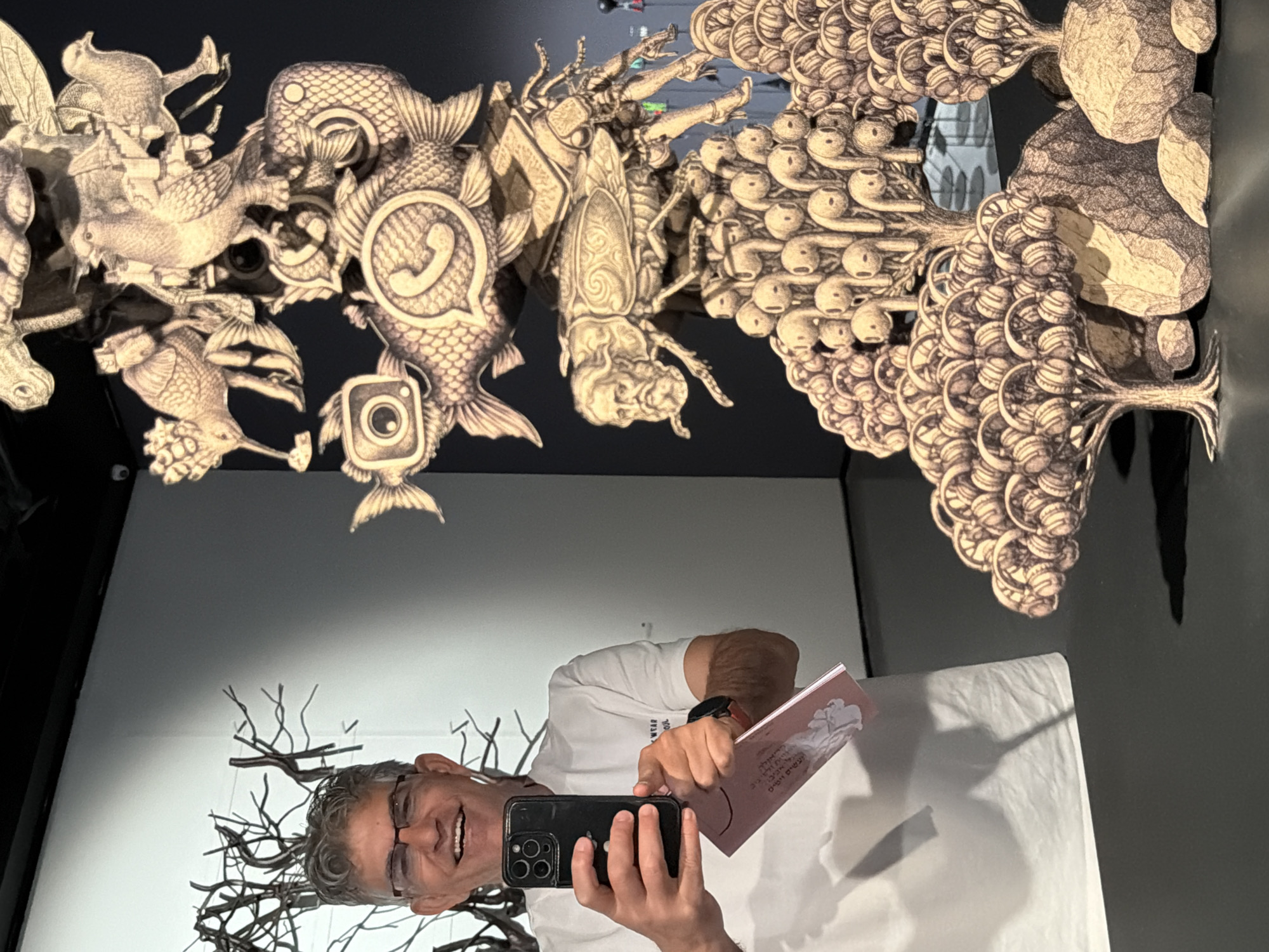 A middle-aged man is taking a selfie in front of an art installation, likely a mirror with mounted sculptures. The sculptures feature surreal creatures and objects rendered in a monotone, textured style. Some elements resemble birds, fish with app icons (like WhatsApp and Instagram), and tree-like structures composed of small, rounded objects. The man has short, graying hair, glasses, and a white t-shirt. He is holding a black smartphone in his left hand and a small, pink booklet in his right. The background consists of white and dark gray walls. The scene is likely within a gallery or museum setting. The exact location in Tel-Aviv, Israel, is not discernible from the image.