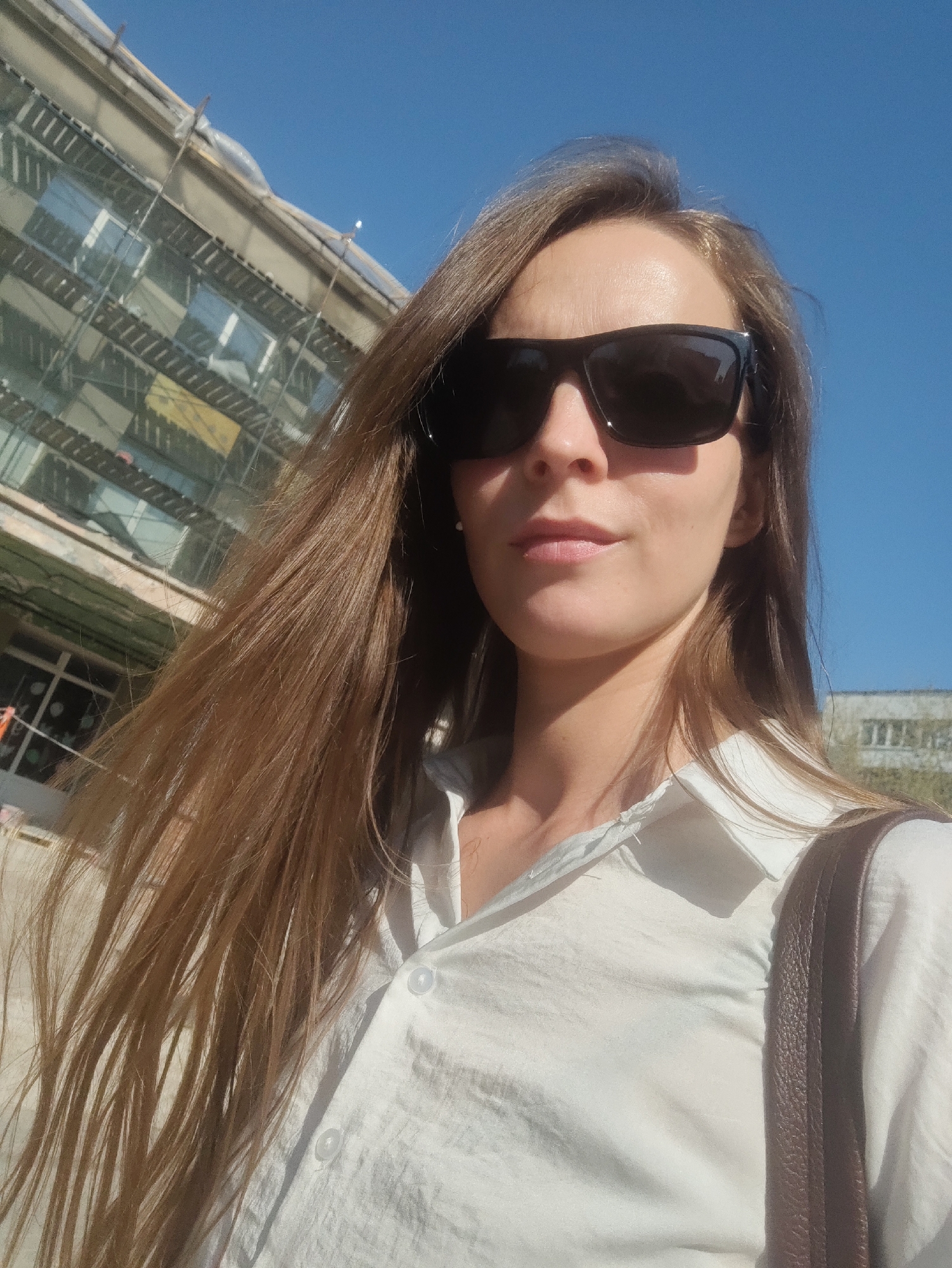A woman with long brown hair and wearing sunglasses and a white button-down shirt is the main subject of the image. She is looking directly at the camera. A brown strap, possibly from a bag, is visible over her shoulder. The background includes a building with scaffolding, partially obscuring its facade, and a clear blue sky. A second building appears smaller and further in the background to the right. The location is the city of Chaikovsky, Russia, indicated by the building's architectural style and urban layout. The time of day appears to be daytime, with bright natural light.