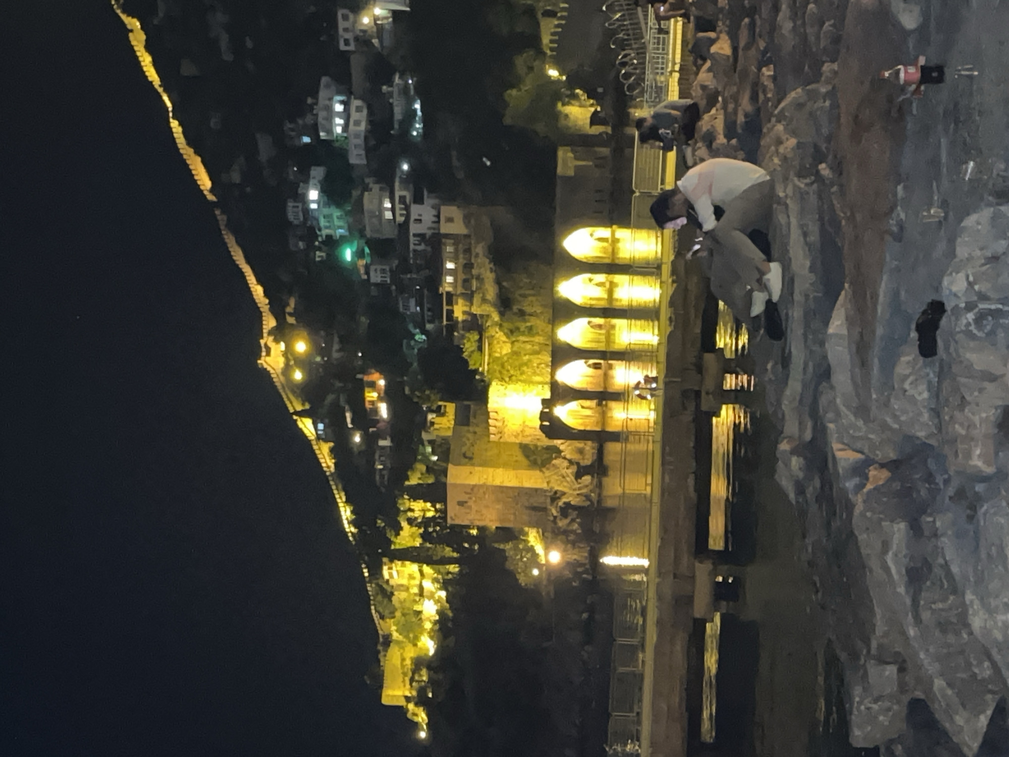 The image captures a vibrant night scene in Alanya, Turkey, showcasing the illuminated historical architecture and coastline. The primary subject is a fortified wall or city rampart, adorned with numerous yellow lights, stretching along a dark hillside on the left, curving towards the horizon. Below the rampart, various buildings with lit windows indicate a populated town or city.
In the midground, a prominent historical structure resembling the Alanya Shipyard (Tersane) or a part of the Red Tower complex stands out. It features several distinctive arched openings, glowing brightly with yellow light, which cast strong, shimmering reflections upon the calm body of water in front of it.
The immediate foreground on the right side reveals a rocky, uneven embankment or shore. Several people are visible here; one individual, wearing a light-colored shirt and dark pants, is seated on the rocks, seemingly engaged with a mobile phone. Another person is partially visible further back.
The scene takes place at night, evident from the dark sky and the reliance on artificial lights to illuminate the structures and landscape. The lights create a warm, inviting glow, contrasting with the dark surroundings. The water acts as a mirror, enhancing the visual spectacle of the illuminated city. No discernible text is visible in the image.