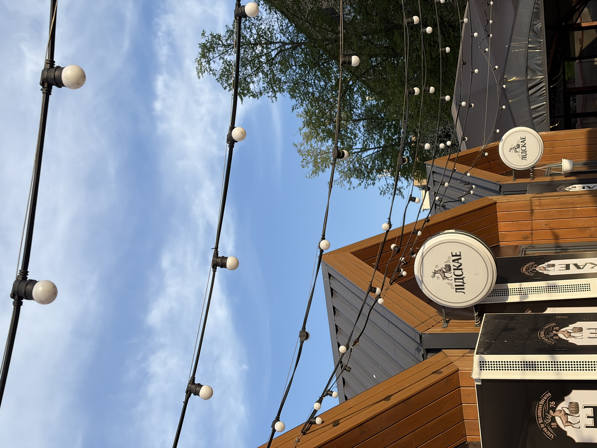 The image captures an outdoor setting, likely a pub or restaurant in Minsk, Belarus, based on the "ЛIДСКАЕ" signage (Lidskoe is a Belarusian beer brand). The scene is shot from a low angle, looking upwards. The sky is visible with scattered clouds. Decorative string lights with globe bulbs are strung across the area. The building features wooden and metal architectural elements. The Lidskoe signs indicate the establishment serves that brand of beer.
