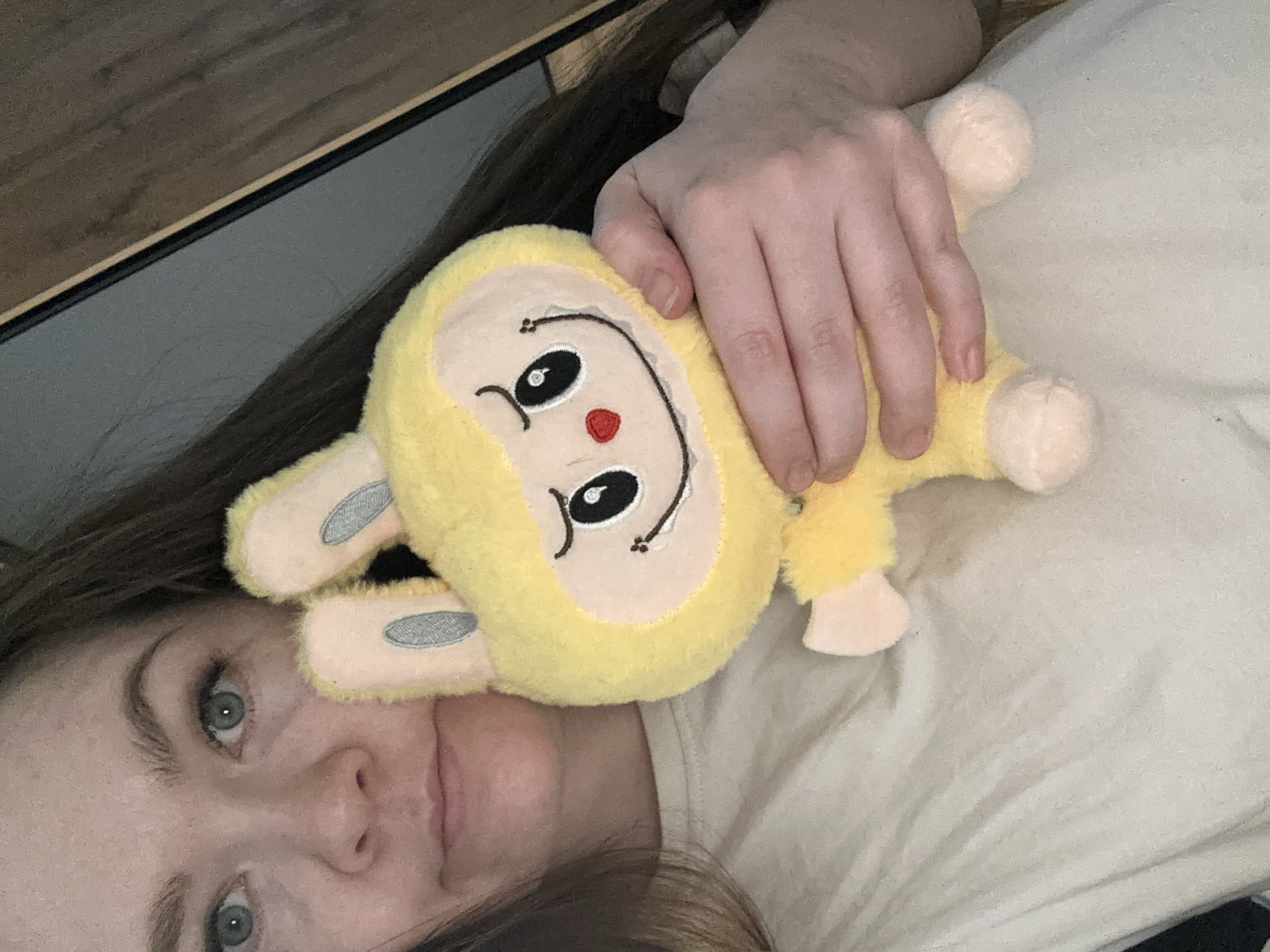 A woman is lying on a bed, holding a yellow stuffed animal. The woman has light skin, green eyes, and brown hair. She is wearing a beige t-shirt. The stuffed animal is a light yellow color with a cream-colored face and ears. It has embroidered facial features. The bed is covered in a beige sheet. A portion of a wooden headboard is visible in the background. There is no information to indicate that the image was taken in Chaikovsky, Russia.