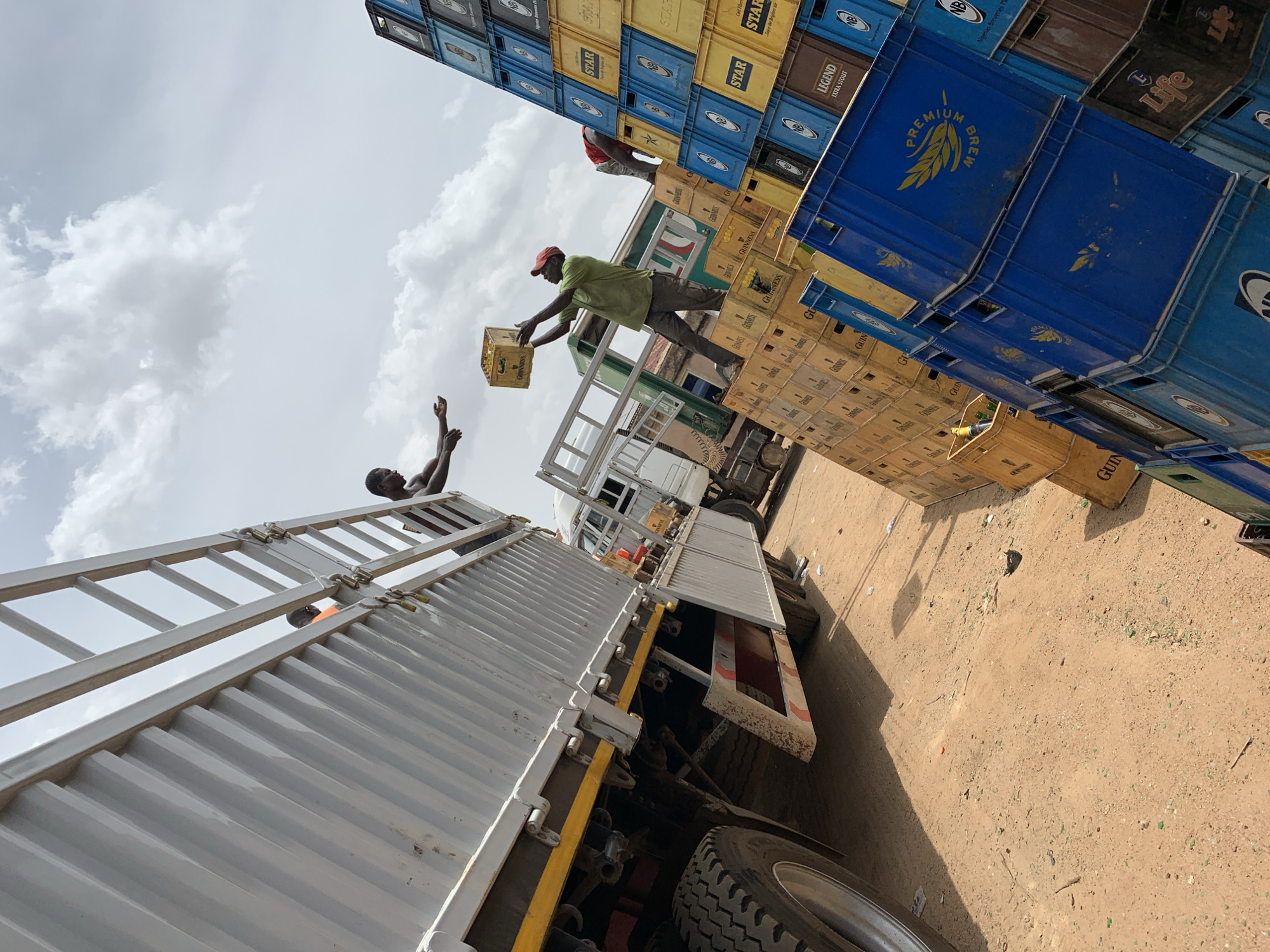 In Bukuru, Nigeria, men are unloading crates, stacked high, from a truck. One man stands atop the crates passing a crate labeled "GUINNESS" down to another man standing on the truck's ladder. Crates of “STAR”, “LEGEND EXTRA STOUT”, “PREMIUM BREW” and “Life” are also visible. The ground appears to be a dirt surface with bits of broken glass scattered about. The sky is partly cloudy.