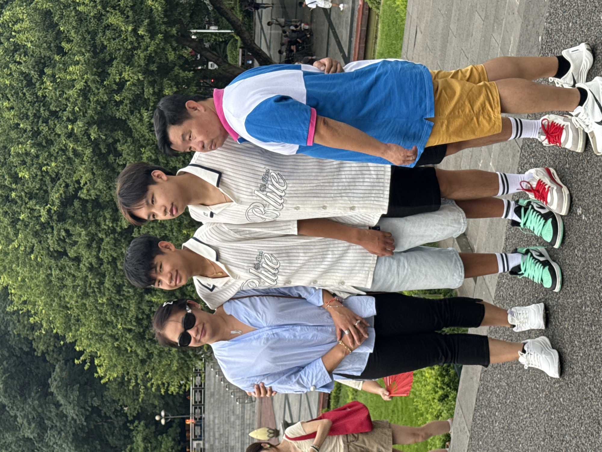 In the city of Yuzhong District, China, a family of four, consisting of two teenage boys, a woman, and a man, poses for a photograph. The woman, on the left, wears a light blue collared shirt, black capri pants, and white sneakers. She has dark hair and wears sunglasses and jewelry. She stands with her left arm around one of the boys. The two boys, standing next to each other, wear matching vertically striped short-sleeved shirts and light-colored shorts. They have dark hair and are dressed in sneakers. The man, on the far right, wears a blue and white polo shirt with pink trim and yellow shorts, paired with white sneakers. He has dark hair and stands with his arm around one of the boys. The family is positioned on a paved walkway in front of a large tree with lush green foliage. In the background, there are stone steps, a lamp post, and other people walking and sitting in the distance. The overall atmosphere appears casual and relaxed, likely a family outing in a park or public space.