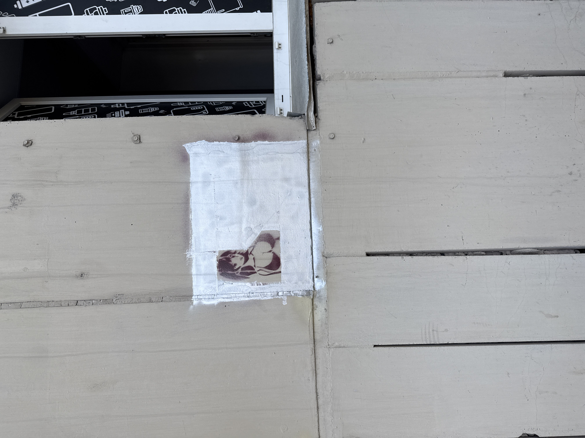 This image shows a section of a light-colored, likely concrete, wall or building exterior. The wall appears to be composed of stacked, rectangular segments with horizontal grooves between them. A recently painted white square covers a section of the wall, partially obscuring a stenciled or printed image of a woman's head and upper body. The location is Unknown, Unknown. A window or opening is visible at the top-left of the image.