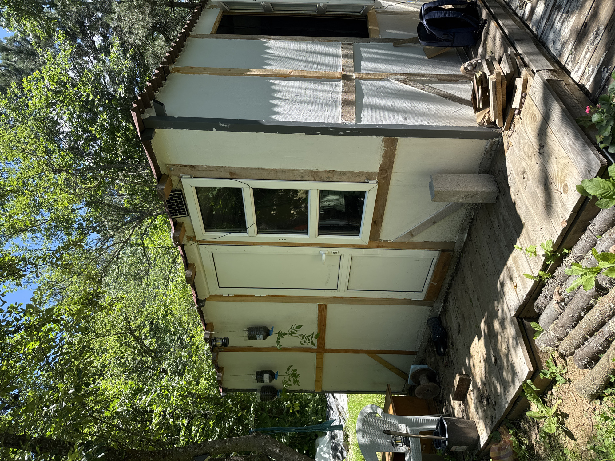The image depicts a small, unfinished structure, possibly a shed or a makeshift dwelling, situated outdoors amidst lush greenery. The building has a rustic appearance, with visible wooden framing and a textured, light-colored exterior finish. A corrugated metal roof with brown trim partially covers the structure.
The front of the building features a white door and two windows. Three plastic bottles, repurposed as planters for small plants, hang from the wall to the left of the door. A solar panel is visible on the roof above the windows.
The ground in front of the structure is a wooden deck, appearing weathered and somewhat uneven. Various objects are scattered on the deck, including a white plastic chair, a black bucket with a tool handle, what looks like a spool of wire, a brick, and a pair of black shoes. Further to the right, near the corner of the building, a stack of wood scraps and a blue backpack are present.
The surrounding environment is characterized by trees and foliage, suggesting a rural or semi-rural setting. The lighting indicates it is daytime, with shadows cast by the structure and trees. The scene appears to be located in Dramsha, Bulgaria, based on the provided information. There are no people visible in the image.