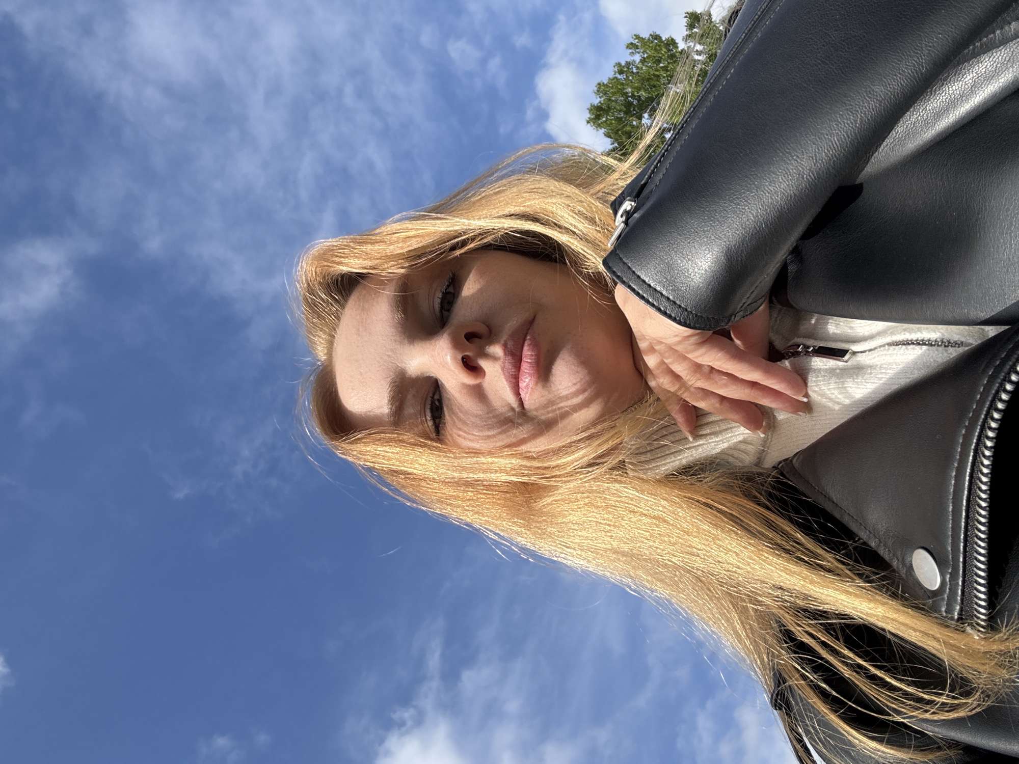 A low-angle shot looking up at a woman with long blonde hair, wearing a black leather jacket over a white sweater. The background is a blue sky with scattered white clouds and a glimpse of green foliage. The woman's face is visible, with a neutral expression. The location is Заволжье, Russia, however the area of the city is not discernable from the image.
