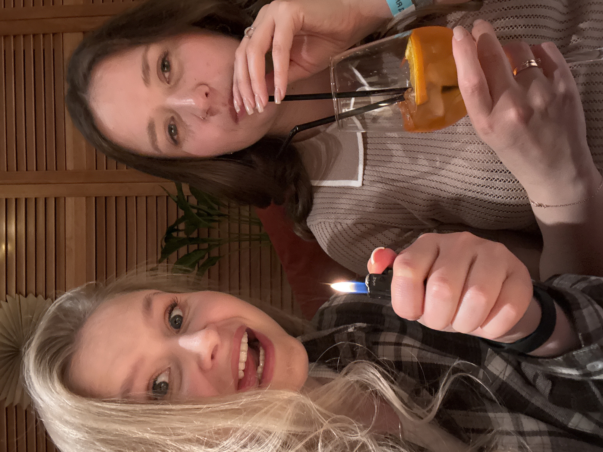 Two women are pictured indoors. The woman in the foreground has blonde hair and is holding a lit lighter in her right hand. She is wearing a gray and white plaid shirt and a black wristband. Her mouth is open in a smile or laugh, revealing her teeth. The woman in the background has brown hair and is drinking from a glass with two straws and a slice of orange. She is wearing a beige sweater with a white collar. A plant is visible behind her, as is a wood-paneled wall. It is not possible to tell where this location is with the given information.