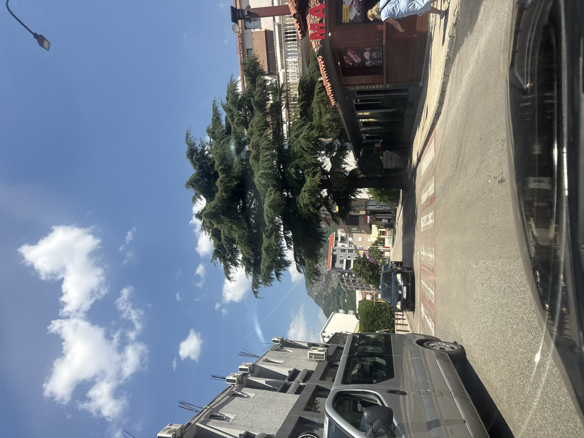 The image captures a street scene in Budva, Montenegro, under a clear blue sky with scattered clouds. A large evergreen tree dominates the mid-ground, its branches spreading widely over the street. To the left, a gray building with multiple air conditioning units is visible, and a silver van is parked in the foreground. Across the street, a small building with a red tiled roof and signage that appears to say "MA" is situated next to a larger building with a white balustrade on its balcony. A black Volkswagen car is driving on the road, approaching a pedestrian crossing marked with red and white stripes. A woman in a light blue dress is walking on the sidewalk to the right of the street. In the background, several other buildings and a mountain are visible. The overall impression is of a sunny day in a developed urban area with a prominent natural element.