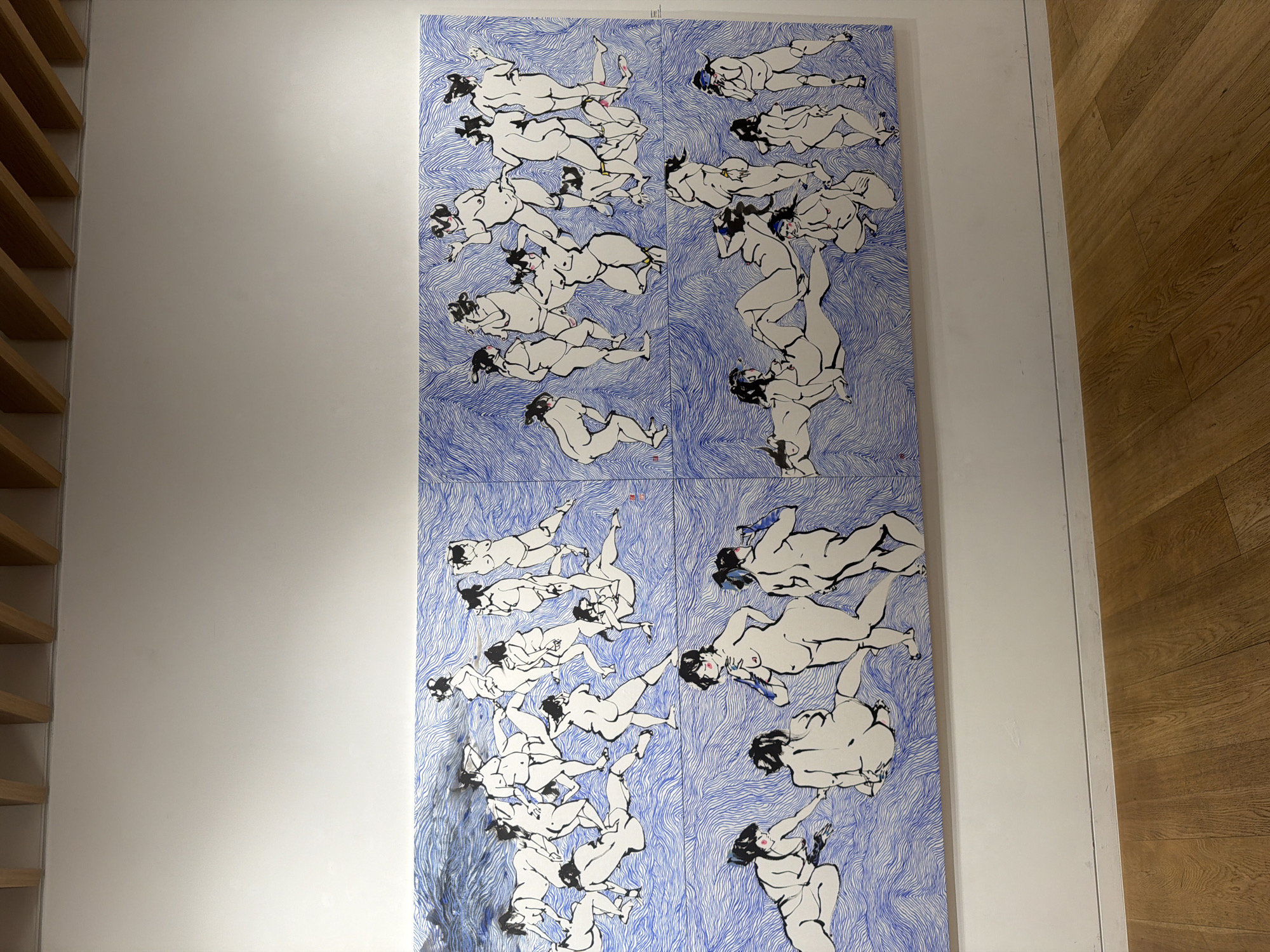 The image depicts a large, multi-panel artwork on a white wall in what appears to be an art gallery or exhibition space. The artwork is a series of ink drawings of nude figures, primarily women, rendered in black ink on a vibrant blue background that mimics flowing water or energetic lines. The figures are stylized and expressive, with varying poses suggesting movement, rest, and interaction. The artwork is divided into four quadrants, with the top two quadrants showing a more dispersed group of figures in dynamic poses, and the bottom two quadrants featuring a more clustered arrangement of figures in relaxed or contemplative positions. The exhibition space features a light-colored wooden floor and a ceiling with exposed wooden beams. The lighting suggests an indoor environment with artificial illumination. There are no people directly visible interacting with the artwork, though the presence of a gallery setting implies a viewership. The overall impression is one of artistic display and cultural engagement. The specific location in Seoul, South Korea cannot be determined from the visual information provided.