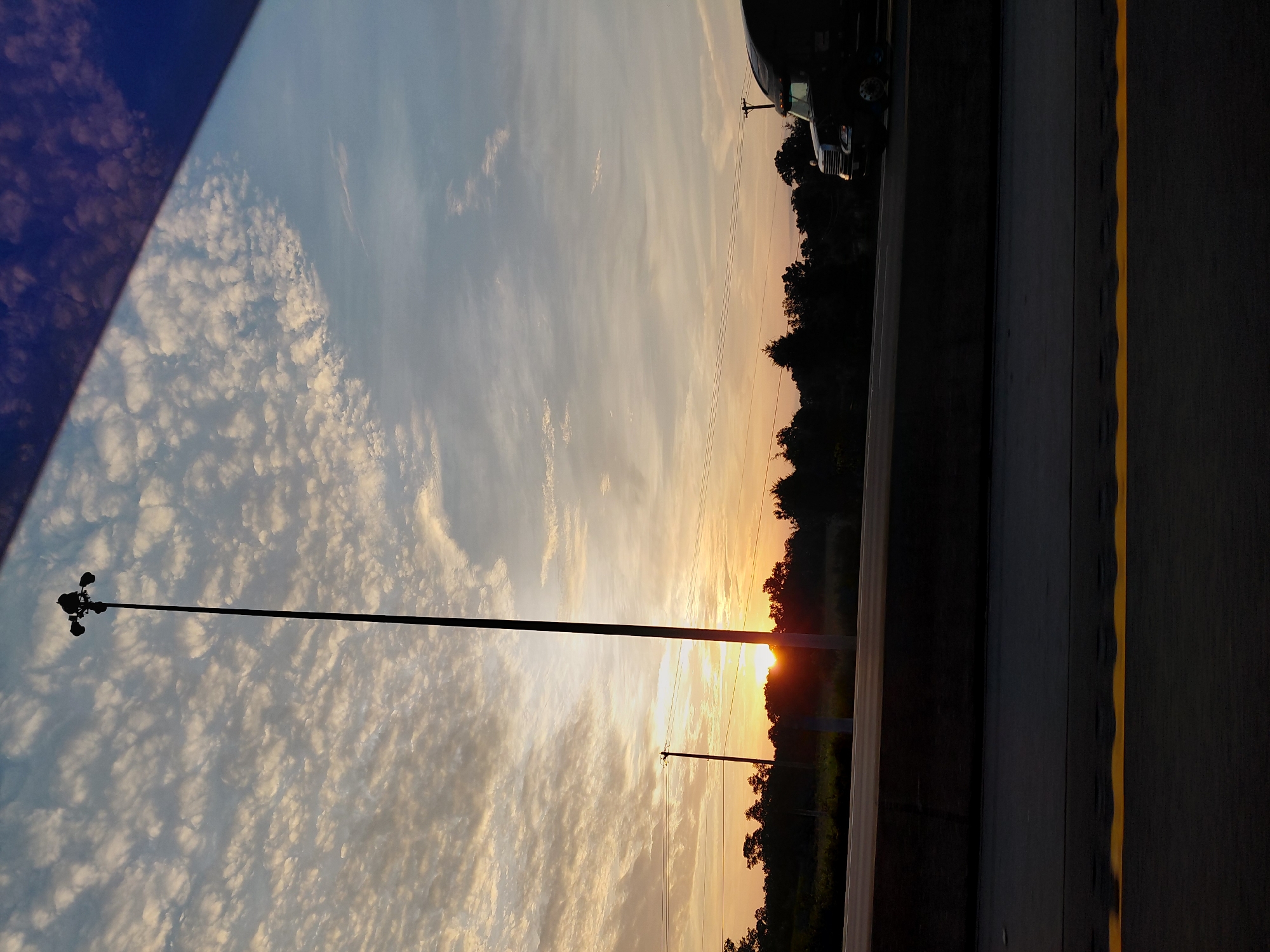 The image captures a scenic view from a vehicle traveling on a highway, likely during sunrise. The sky is adorned with a soft blend of blues and oranges, with scattered, wispy clouds creating a textured effect. A tall, dark lamppost stands prominently in the center left of the frame, silhouetted against the brightening sky. In the distance, a line of trees forms a dark horizon, with the sun just beginning to peek over them. A semi-truck is visible on the right side of the highway, also silhouetted and partially obscuring the tree line. The foreground shows a section of the highway asphalt with a distinct yellow line marking the lane. There are no visible people in the image. The scene suggests a moment of natural beauty encountered during travel, with the urban environment implied by the highway and lamppost.