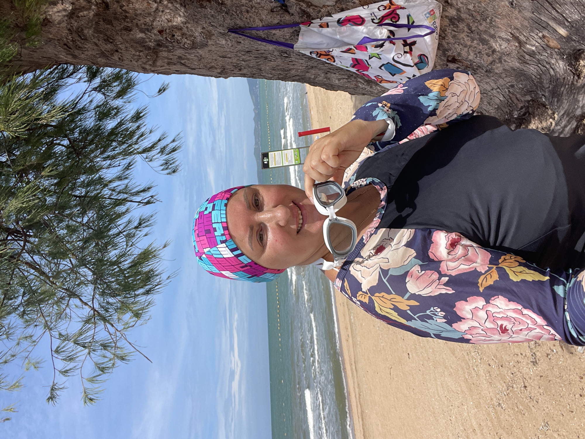 A woman, wearing a colorful swim cap and a long-sleeved rash guard with a floral print, stands on a sandy beach in Pattaya City, Thailand. She is holding a pair of white swimming goggles up to her face. Behind her, the turquoise ocean stretches out to the horizon, with a line of yellow buoys marking a boundary. A large tree trunk is to her right, with a tote bag hanging from it. The sky is blue with scattered white clouds. The overall atmosphere is relaxed and summery.