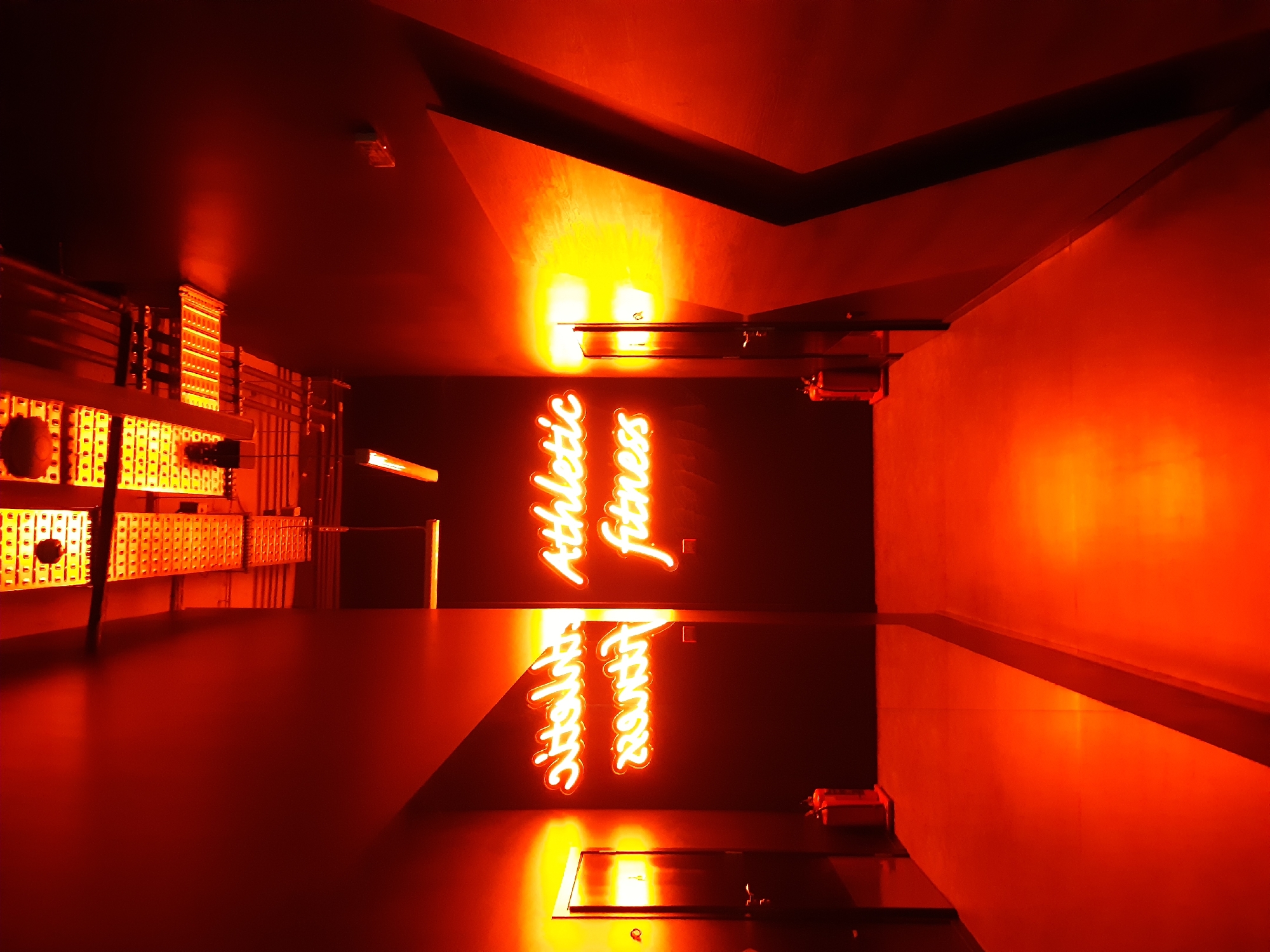 The image is taken indoors in a space with red lighting, possibly a gym. The sign "Athletic Fitness" is visible on the back wall. There is a reflection of the sign on a surface in the foreground. To the left, there is a metal rack system with visible holes, possibly a weight rack. The location is Plovdiv, Bulgaria, as inferred from a search.