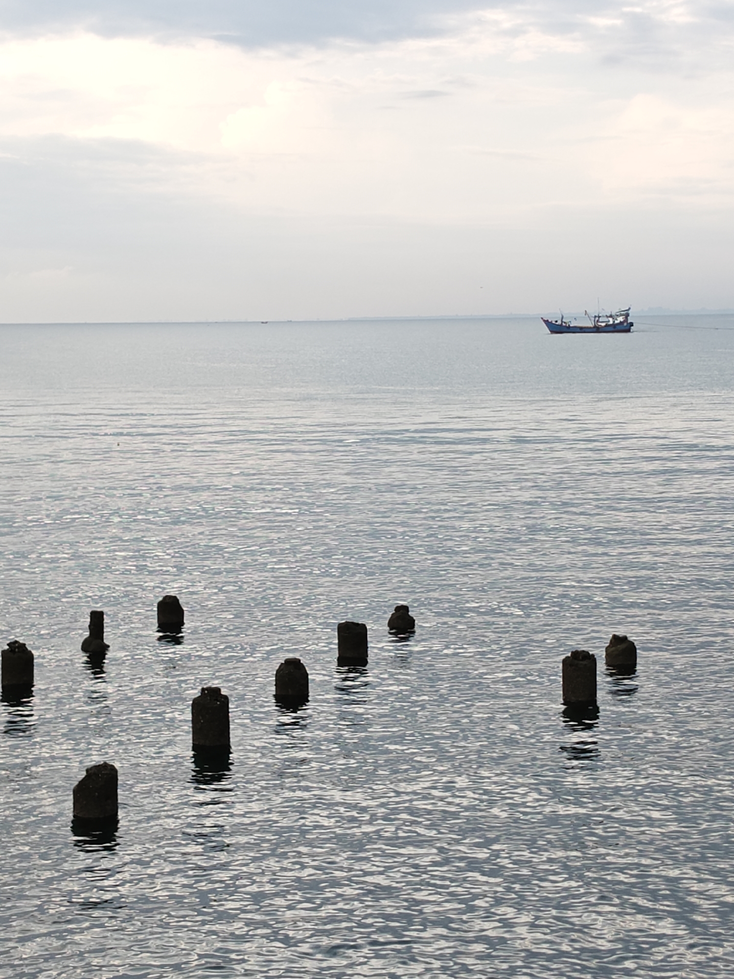 A serene view of the waters off Lhokseumawe, Indonesia, captures the tranquil beauty of the sea under a cloudy sky. In the foreground, several weathered, dark-colored posts emerge from the gentle waves, their rough textures contrasting with the smooth surface of the water. These posts, remnants of a bygone structure, stand as solitary sentinels, their bases submerged and their tops exposed.
In the middle ground, the vast expanse of the sea stretches to the horizon, reflecting the soft, muted tones of the sky. The water's surface is a tapestry of subtle ripples, creating a shimmering effect that hints at a light breeze.
Further out, a solitary blue fishing boat navigates the calm waters. The boat, with its distinct silhouette, is engaged in its daily work, a testament to the maritime livelihood of Lhokseumawe. Its presence adds a touch of life and activity to the otherwise peaceful scene.
The sky above is a canvas of overcast clouds, their soft hues creating a diffused light that bathes the entire scene in a gentle, ethereal glow. The horizon is a subtle line where the sea meets the sky, blurred by the atmospheric conditions. In the far distance, hints of land or structures on the coastline can be vaguely perceived, adding a sense of depth and perspective to the image.
The overall atmosphere of the image is one of quiet contemplation and natural beauty, a snapshot of the coastal life in Lhokseumawe, Indonesia.