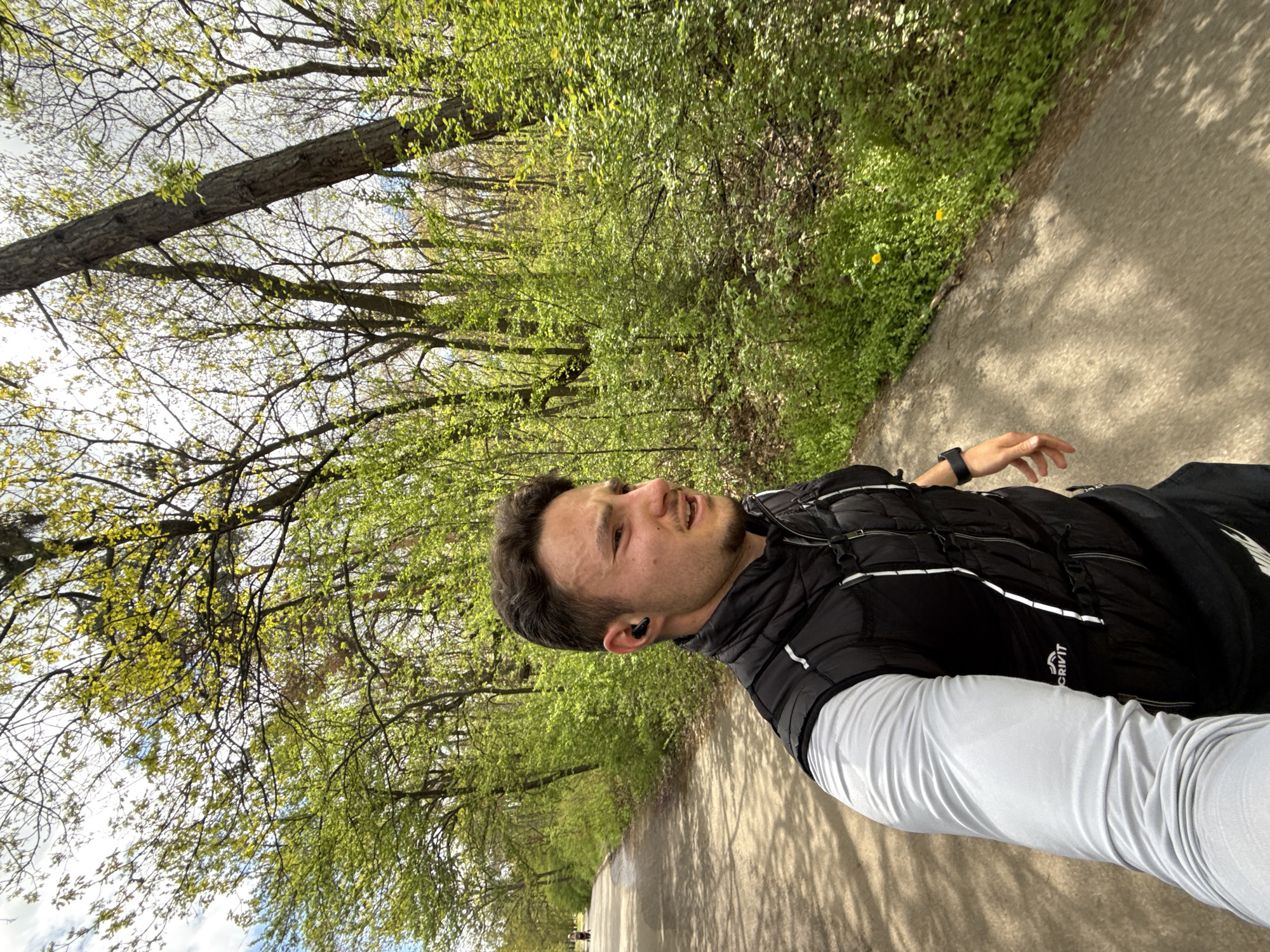 A selfie-style portrait captures a man outdoors on a paved path in Sofia, Bulgaria, likely a park or wooded area during springtime. He is the primary subject, positioned in the foreground, facing slightly right with his head tilted. He has short dark hair and a beard, and wears a black quilted vest with white reflective accents over a light grey long-sleeved athletic shirt and dark pants. A black smartwatch is on his left wrist, and a black wireless earbud is in his right ear. His mouth is slightly open, suggesting exertion or breathing deeply.
The background features a lush natural environment with numerous trees boasting fresh, bright green leaves, indicating spring foliage. The paved path stretches into the distance, with dappled sunlight and shadows from the overhead canopy. Green ground cover with small yellow flowers lines the side of the path. In the far distance, faint figures of other people can be seen on the path, hinting at a public recreational area. The sky, visible through the tree branches, is bright with some clouds. The text "CRIVIT" is visible on his black vest.