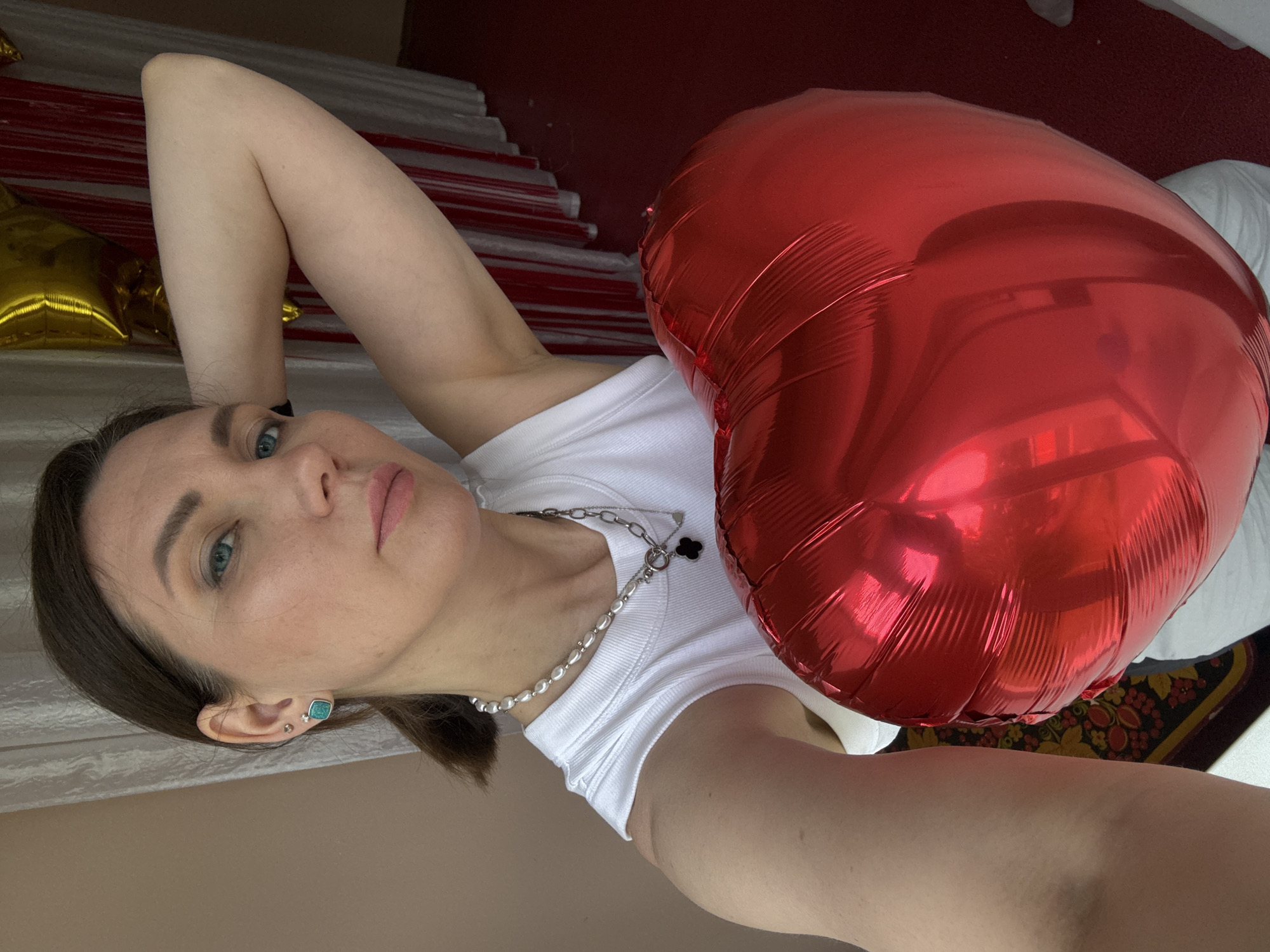 A selfie shows a light-skinned woman with brown hair, blue eyes, and light pink lips lying on a surface. She is wearing a white tank top and a silver chain necklace. In her hand is a large red heart-shaped balloon. In the background, there is a white pillow and a red textured surface. The photo appears to be taken indoors in Chaikovsky, Russia.