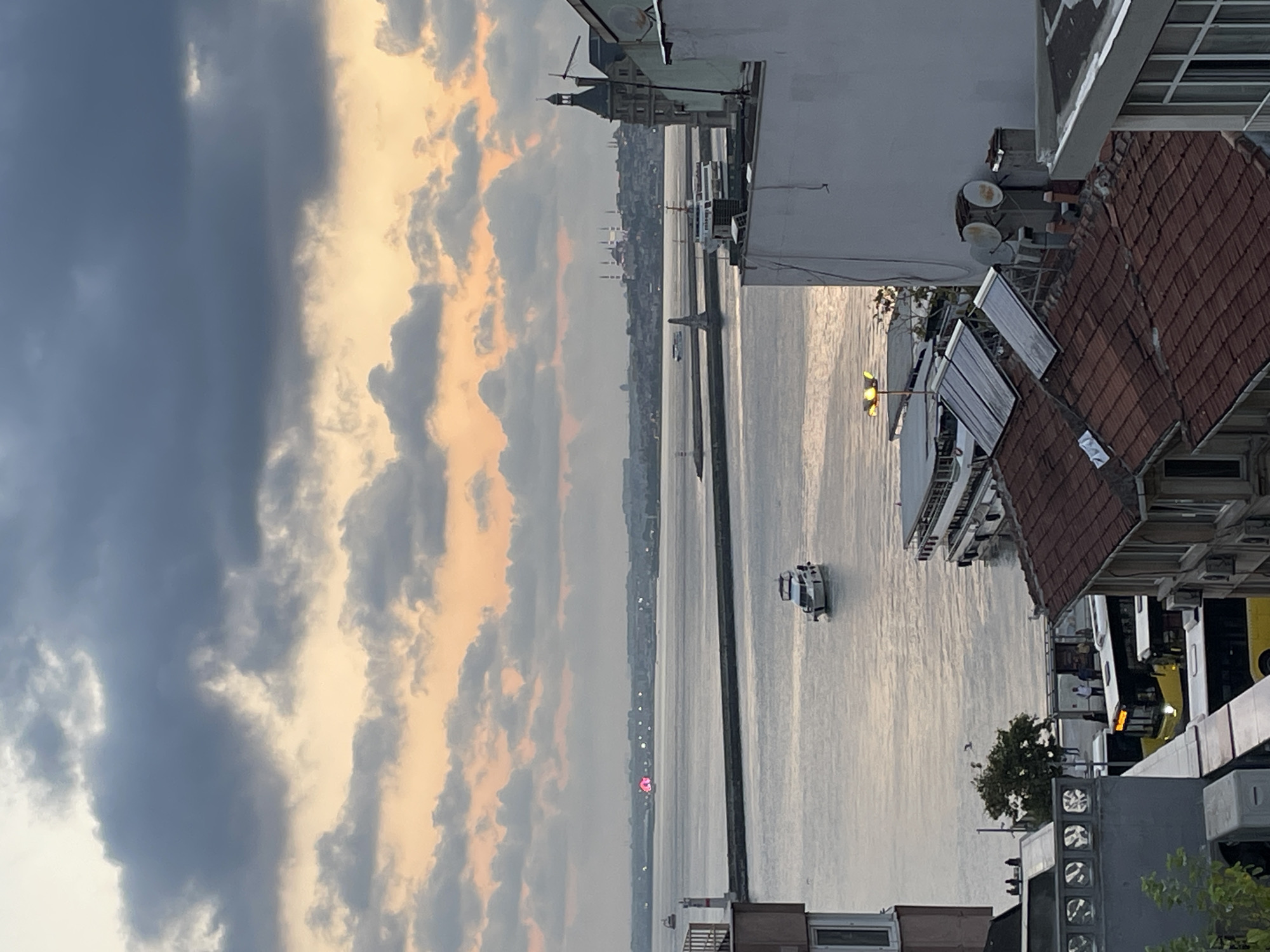 The image captures a dusky scene overlooking the Bosphorus Strait, likely from Kadıköy, Turkey, given the presence of ferry terminals and distinctive architecture. The sky is dominated by dramatic, layered clouds tinged with the orange and yellow hues of sunset. In the foreground, rooftops of buildings with red tiles are visible, some featuring solar panels and satellite dishes. Several yellow and white buses are parked along a waterfront promenade, where a few silhouetted figures can be seen.
The water of the strait is calm, reflecting the muted sky. Several boats are present: a white motor yacht navigates the water, and a larger, multi-deck ferry is docked or moving near the shore. Further out in the strait, a breakwater extends into the water, marked by a lighthouse. In the distance, a city skyline is discernible, with iconic domed structures suggesting historical buildings like Hagia Sophia and the Blue Mosque. A Ferris wheel, illuminated with pink lights, is visible on the far left horizon. The overall atmosphere is one of a serene twilight, with the bustling activity of the city implied by the presence of transportation and residential buildings.