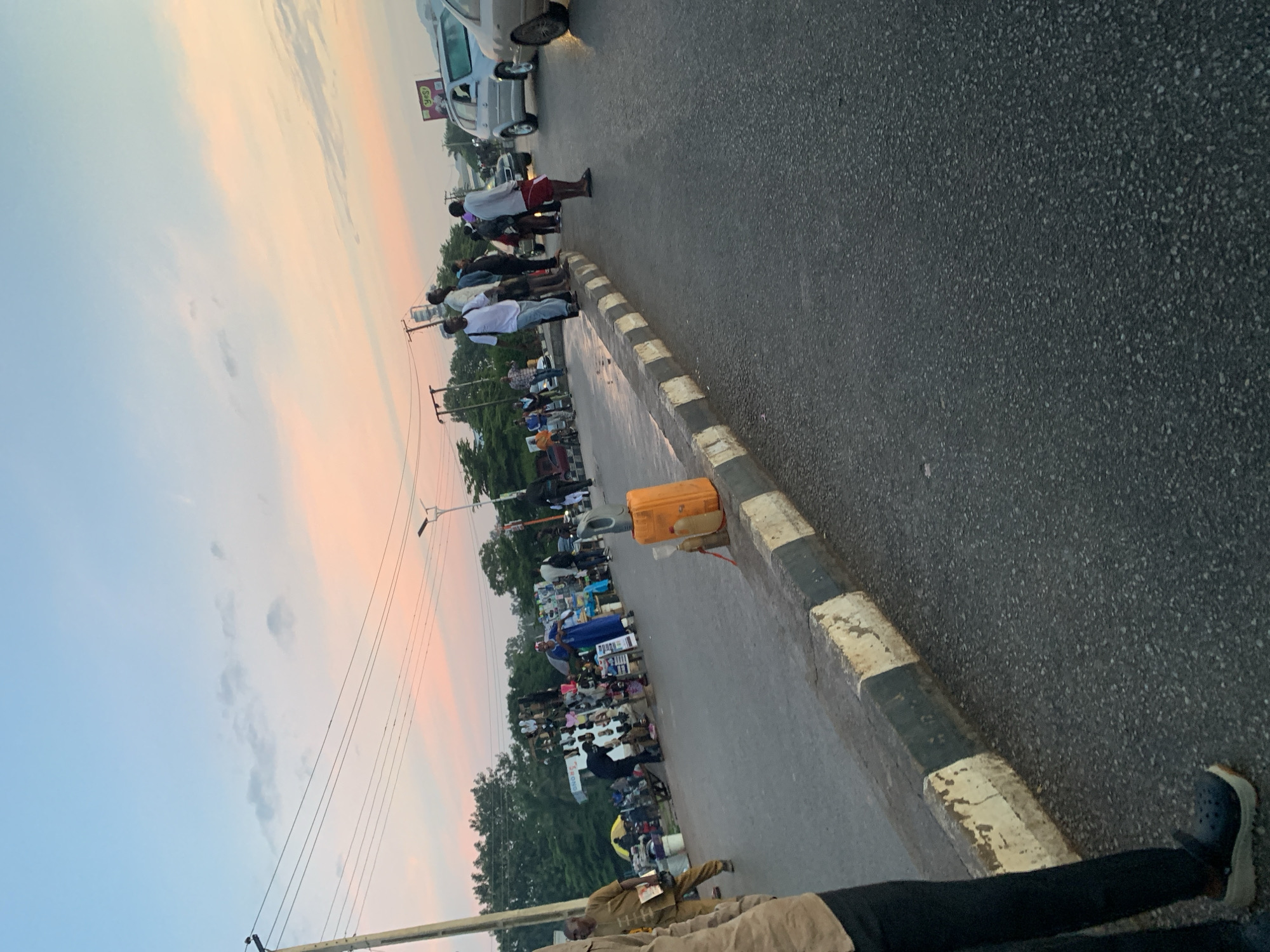This image captures a street scene in Jos, Nigeria, during what appears to be either sunrise or sunset, given the colorful sky. The road is asphalt and appears to be a main thoroughfare, with a raised concrete curb separating the road from a sidewalk or pedestrian area. Several people are present on the sidewalk and on the edge of the road.
On the left side of the image, near a utility pole, a street vendor is displaying footwear on a rack. A person in a brown outfit is visible behind the vendor. Further down the sidewalk, there are more stalls with various items for sale, including what looks like clothing. A person wearing a blue traditional garment is standing near these stalls.
On the right side of the image, a group of people are standing on the edge of the raised curb, some of whom appear to be waiting or observing the traffic. Several vehicles are visible on the road, including a silver SUV and a lighter-colored car. An orange plastic container, possibly a fuel jerrycan, and some bottles are placed on the ground near the curb in the foreground. The background shows a line of trees and some buildings. The sky is a gradient of pale blue and soft pinks, indicating the time of day.