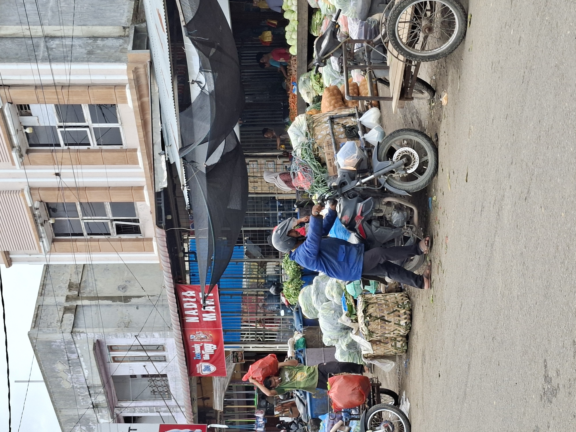 A street market scene in Lhokseumawe, Indonesia, is depicted on an overcast day. In the foreground, a man wearing a blue jacket and a helmet is pushing a motorcycle to the right. To his left, another man in a green t-shirt carries a large red plastic bag over his shoulder. Behind them, a variety of produce is displayed, including large sacks of vegetables and piles of leafy greens, some of which are contained in woven baskets. A red banner with "NADIA MART" written on it hangs above the market stalls. In the background, market stalls are set up under a dark tarp, with several people visible, including a woman in a hijab. The ground is paved, with scattered debris. The buildings surrounding the market appear to be two-story structures, some with faded paint. The overall impression is of a bustling outdoor market.