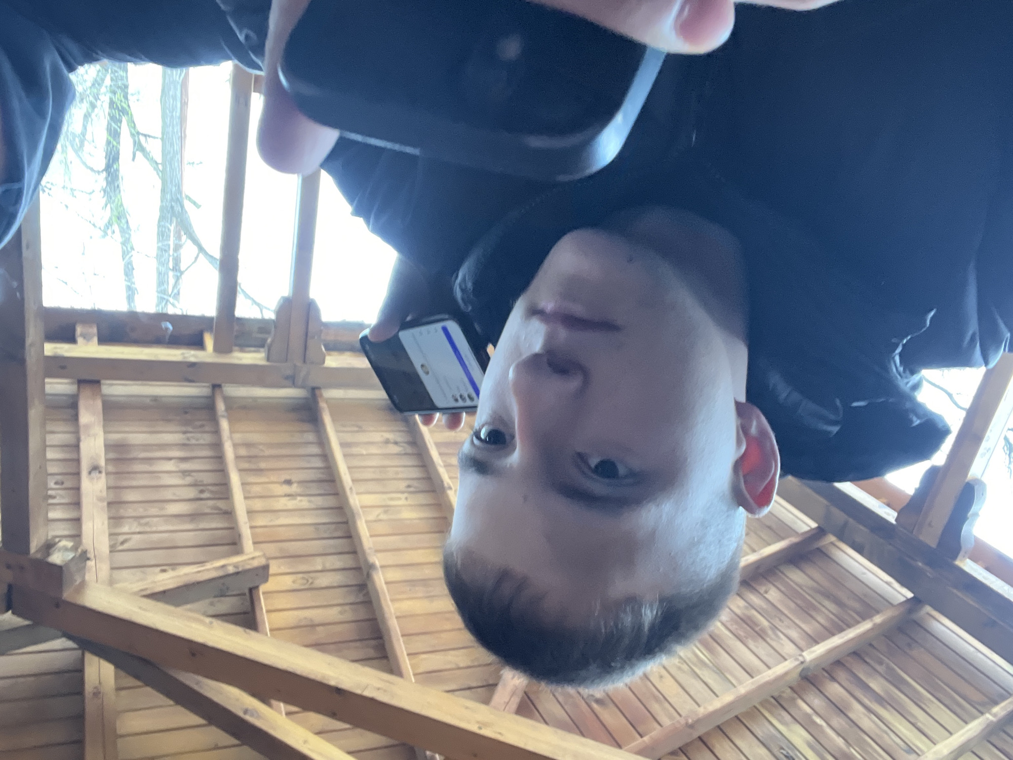 A man is taking a selfie inside a wooden structure. He holds a phone in his right hand, slightly above his head. The structure appears to be a gazebo or a small shed, with wooden beams and planks visible in the roof and walls. Through the gaps in the structure, trees can be seen in the background. The location is unknown and there is no indication to relate the photo to Asenovgrad, Bulgaria.