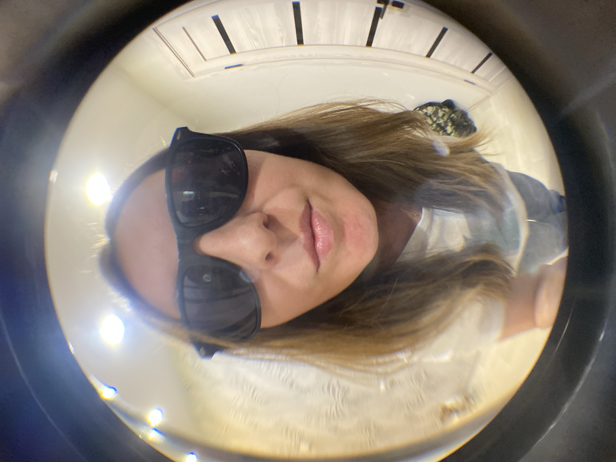 A person is captured in a highly distorted, circular image, likely taken through a fisheye lens or reflected in a spherical mirror. The primary subject is a person, appearing to be an adult female with shoulder-length brown hair, wearing black sunglasses and a light-colored top. Her face is turned slightly, with a subtle smile visible beneath the sunglasses.
The image is heavily distorted, with the subject's head and upper torso filling much of the circular frame. The background is light-colored, possibly a wall or ceiling, with several vertical black lines visible, which could be part of a large clock face or decorative architectural features. The entire scene is framed by a dark, circular border, emphasizing the unique perspective. Bright circular reflections, possibly from overhead lights, are visible in the lower portion of the frame, indicating an indoor setting.
No specific activity beyond posing for the photograph is evident. The image does not provide cues for time of day or weather. While the location context of Noviy, Russia, is provided, there are no visual details within the image to specifically identify it as such. No visible text is present.