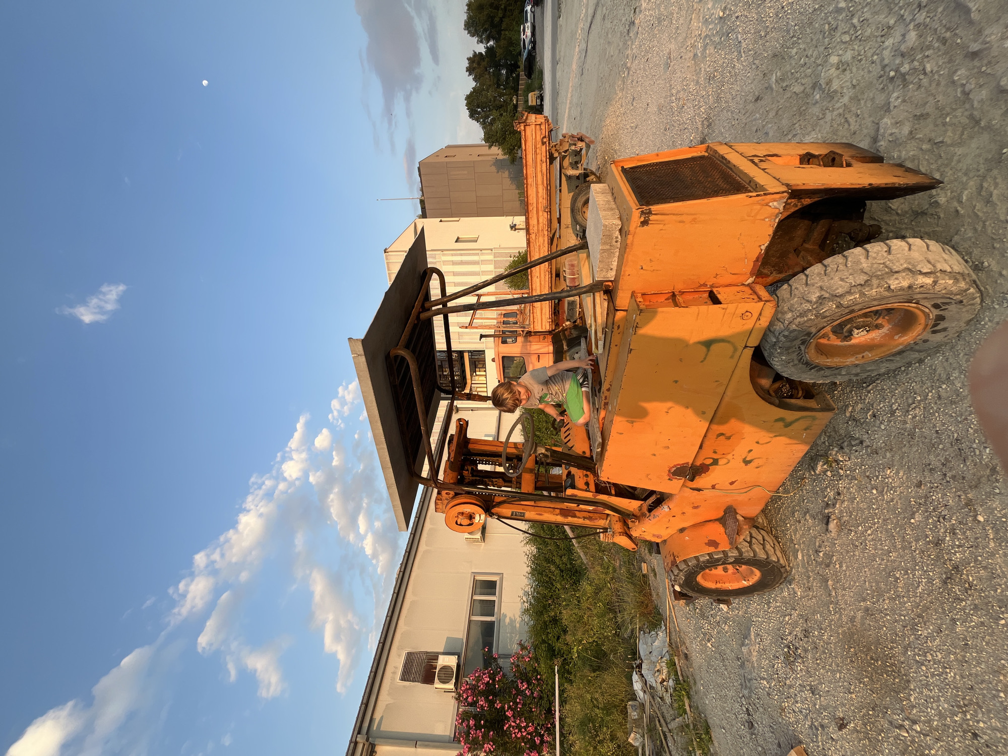 The image captures a young boy sitting in the driver's seat of an old, orange forklift parked on a gravelly lot in Pobrdje, Montenegro. The forklift is weathered and shows signs of rust. A building with a white facade and visible air conditioning units stands behind the forklift, accompanied by a bush with pink flowers. Beyond the immediate foreground, a taller building and some parked cars are visible, suggesting an urban or industrial setting. The sky is partially clear with scattered clouds and a faint moon visible in the upper left corner. The lighting suggests it is late afternoon or early evening. The boy is wearing a t-shirt and shorts, and he appears to be engaged in the act of playing or pretending to operate the machinery.