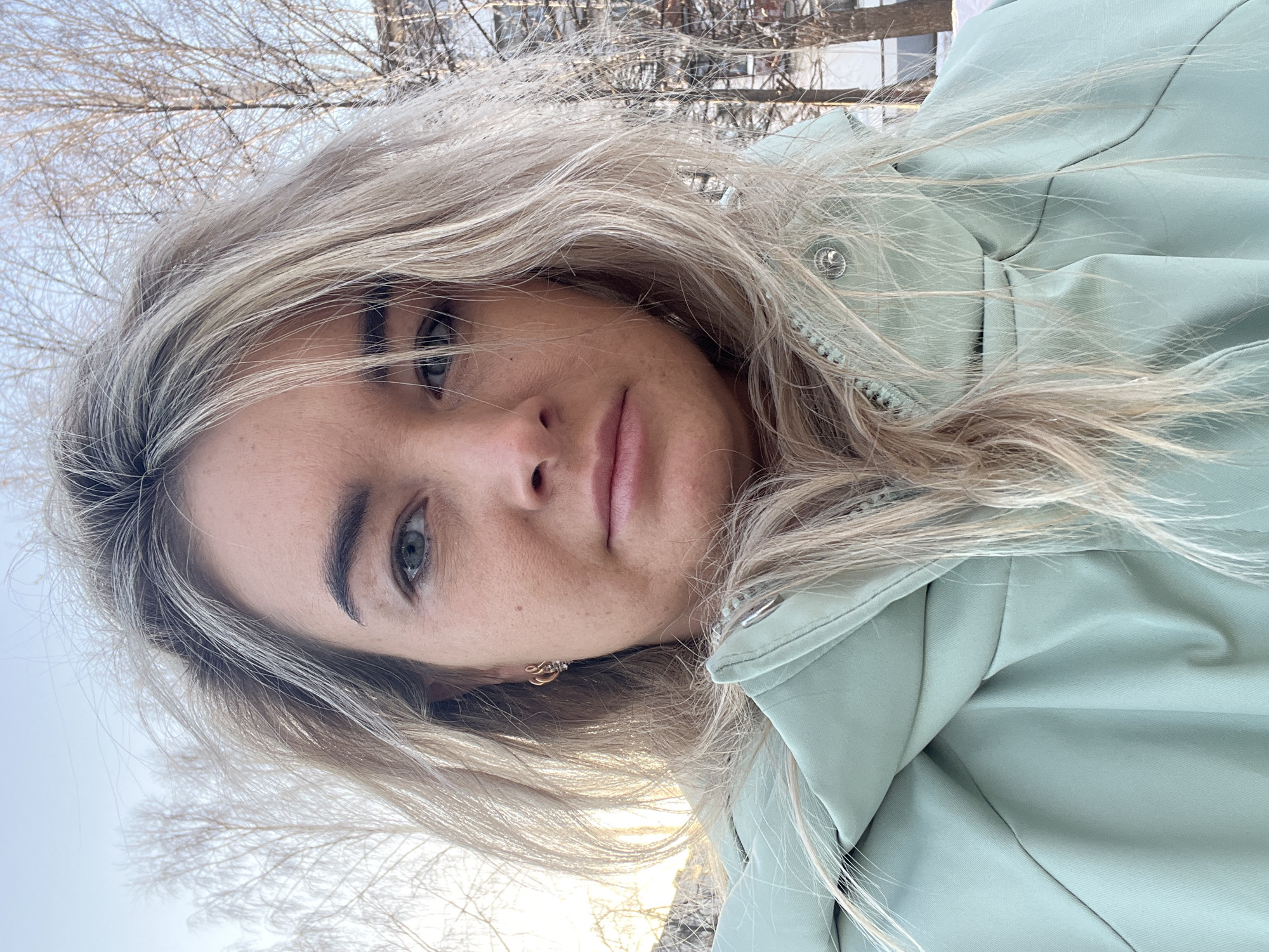 A medium shot shows a young woman with light complexion and blonde hair, wearing a light green jacket, looking towards the camera. Bare tree branches are visible in the blurred background, suggestive of a wintry or early spring setting. Given the prompt's requirement to place the image in Chaikovsky, Russia, one might presume she is in an outdoor area within this city, possibly a park or residential zone adjacent to open spaces or trees. Buildings are also faintly visible behind the branches. She has light eyes, possibly blue or green. The lighting indicates it could be daytime, though the time of day isn't definitively clear.