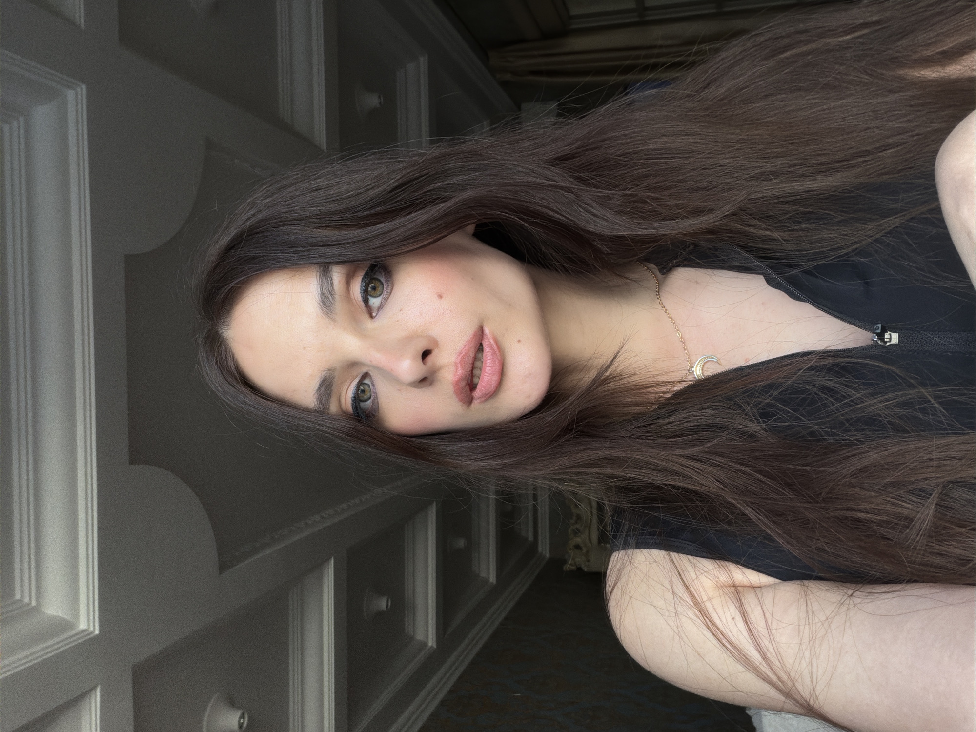 This is a close-up, vertically oriented selfie of a young woman with long, dark brown hair and light skin. She is wearing a black top and a delicate gold necklace with a crescent moon pendant. Her eyes are light green, and she is wearing makeup, including eyeliner and mascara, and her lips are subtly glossed. She has a small mole on her cheek.
The background appears to be an indoor setting. To the left of the woman, there is a wall with decorative paneling and molding, suggesting a home interior. The lighting suggests it might be daytime, possibly with natural light filtering in. There is no visible text in the image. The woman's expression is neutral, and the overall mood is calm and casual. The context provided indicates the location is Ufa, Russia.