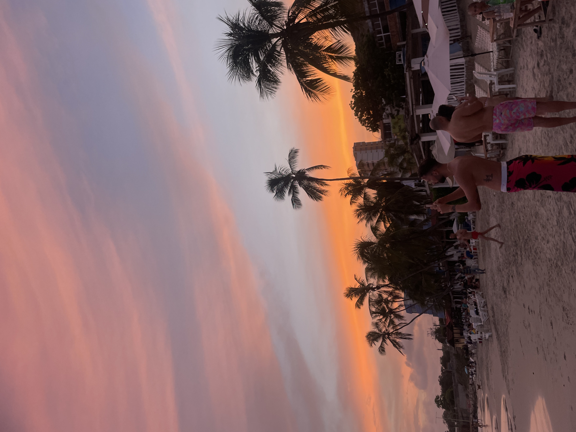 This is a vertical, eye-level shot of a beach at sunset in Pampatar, Venezuela. The sky displays a gradient of pastel colors, transitioning from a soft pink at the top to a vibrant orange and yellow near the horizon. Silhouetted palm trees with full fronds punctuate the scene, with their tops reaching into the colorful sky.
In the foreground, a sandy beach stretches across the frame. The tide is out, revealing wet sand near the water's edge. Several people are visible on the beach. On the left, a man with tattoos is taking a picture with his phone, wearing a bright pink sarong with a floral pattern. To his right, another man is seen from behind, wearing brightly colored floral swim trunks. Further back, a few other individuals are scattered across the beach.
Behind the beach, a line of low-rise buildings, possibly hotels or restaurants, are visible, with some featuring white awnings and umbrellas. The architecture suggests a tropical coastal town. In the distance, a multi-story building stands out against the horizon. The overall atmosphere is relaxed and serene, characteristic of a popular tourist destination during golden hour.