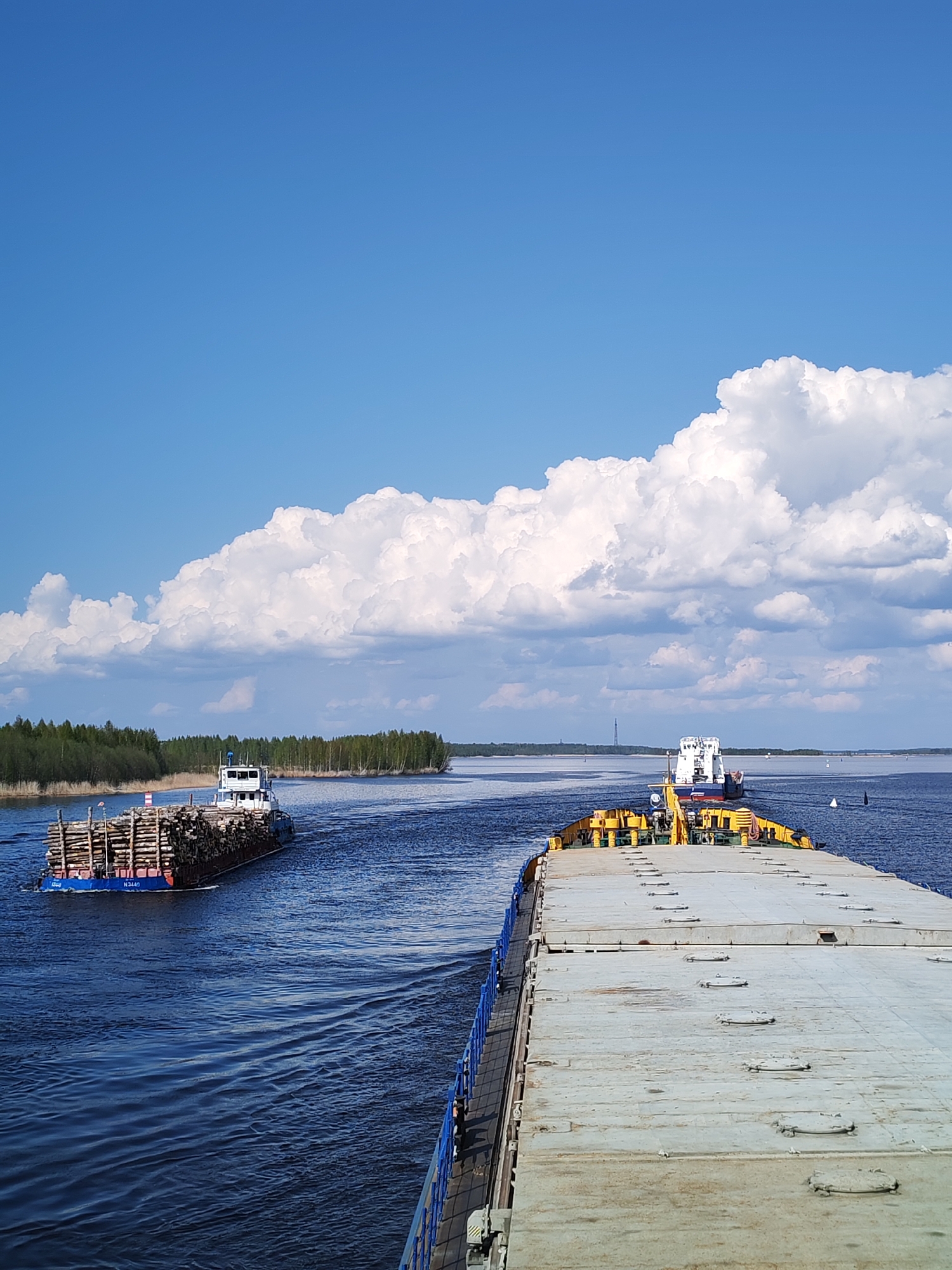 The image shows a wide river with two barges on it under a partially cloudy sky. One barge in the distance, carrying a load of logs, is headed away from the viewer. In the foreground is the deck of a larger, flat-topped barge with yellow and gray panels. The river flows through a green, forested area. The location is identified as Popovo, Russia.