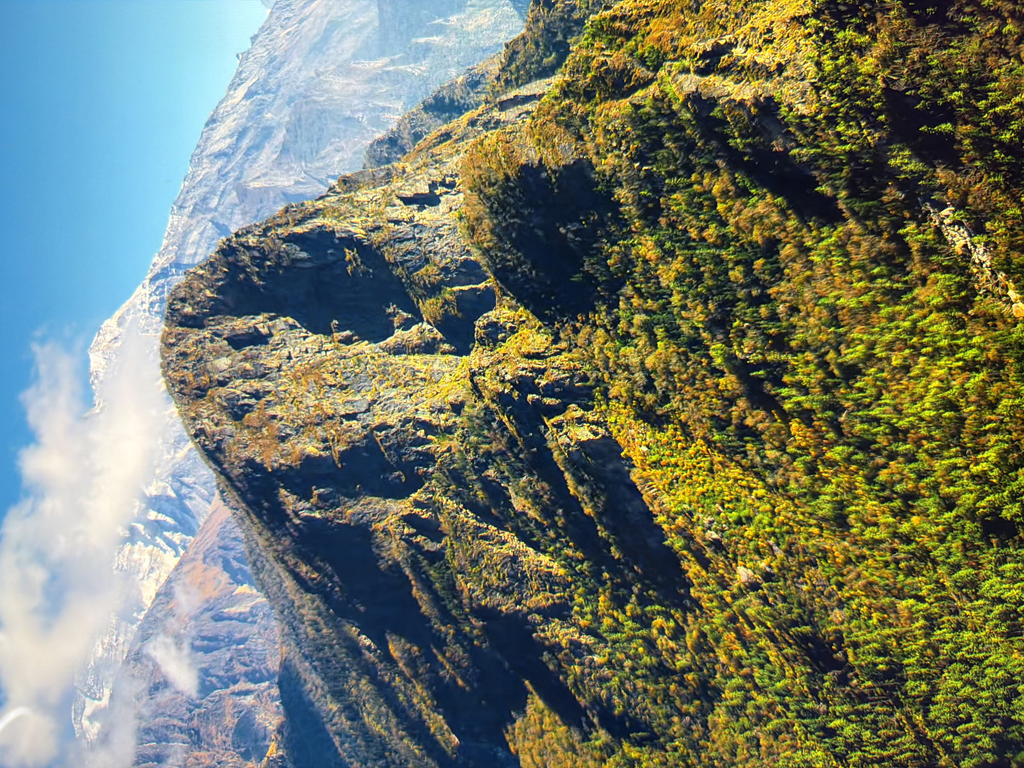 The image is a scenic, aerial view of a mountainous landscape under a bright blue sky. In the foreground, a rugged, rocky mountain peak is prominent, with steep slopes covered in vibrant green and yellow foliage, indicative of autumn. The textured rock faces of this mountain are illuminated by sunlight, casting deep shadows. Behind this peak, further mountain ranges recede into the distance. The furthest mountains are snow-capped and appear hazy, blending into the atmosphere. Patches of white clouds are visible in the sky, particularly around the snow-capped peaks. The overall impression is one of natural grandeur and rugged beauty. The image does not depict any urban environments, people, or man-made objects and therefore cannot be associated with the city of Kyiv, Ukraine.