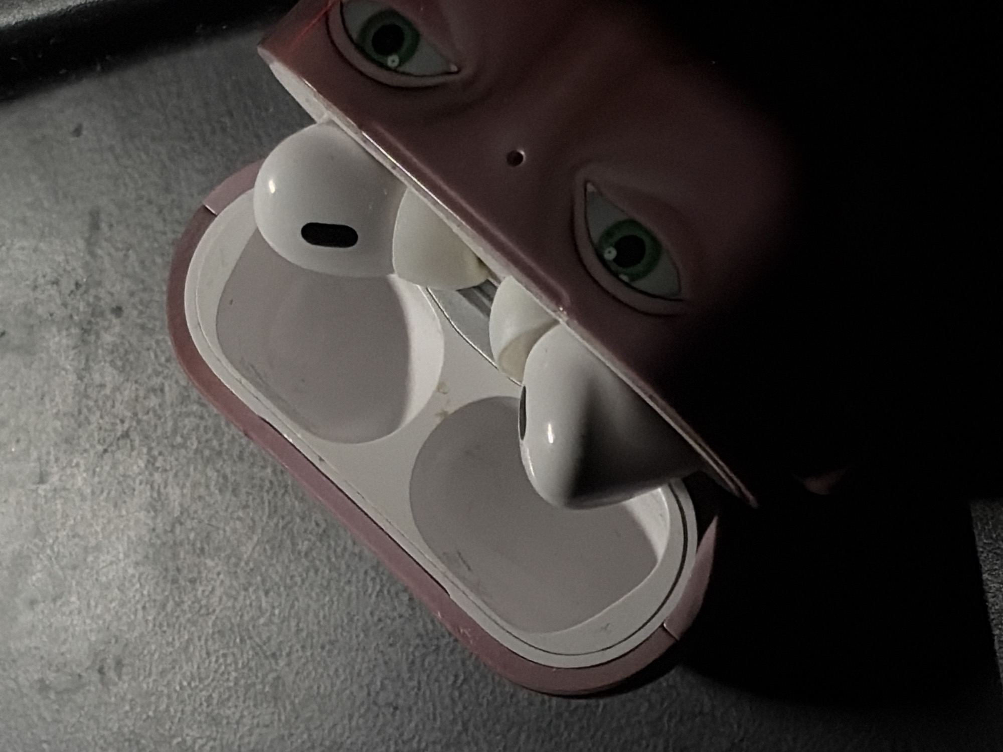 A pair of white wireless earbuds, resembling Apple AirPods Pro, are depicted within their open charging case. The case is enclosed in a pink, silicone-like protective cover, featuring two stylized green eyes with black pupils on one side, and a small circular indentation located centrally above them. Both earbuds are partially visible inside the white interior of the case, which appears somewhat worn or smudged. The assembly rests on a dark, textured surface, consistent with stone or asphalt, under low illumination, creating soft shadows. No discernible features within the image indicate a specific location in the city of Moscow, Russia.