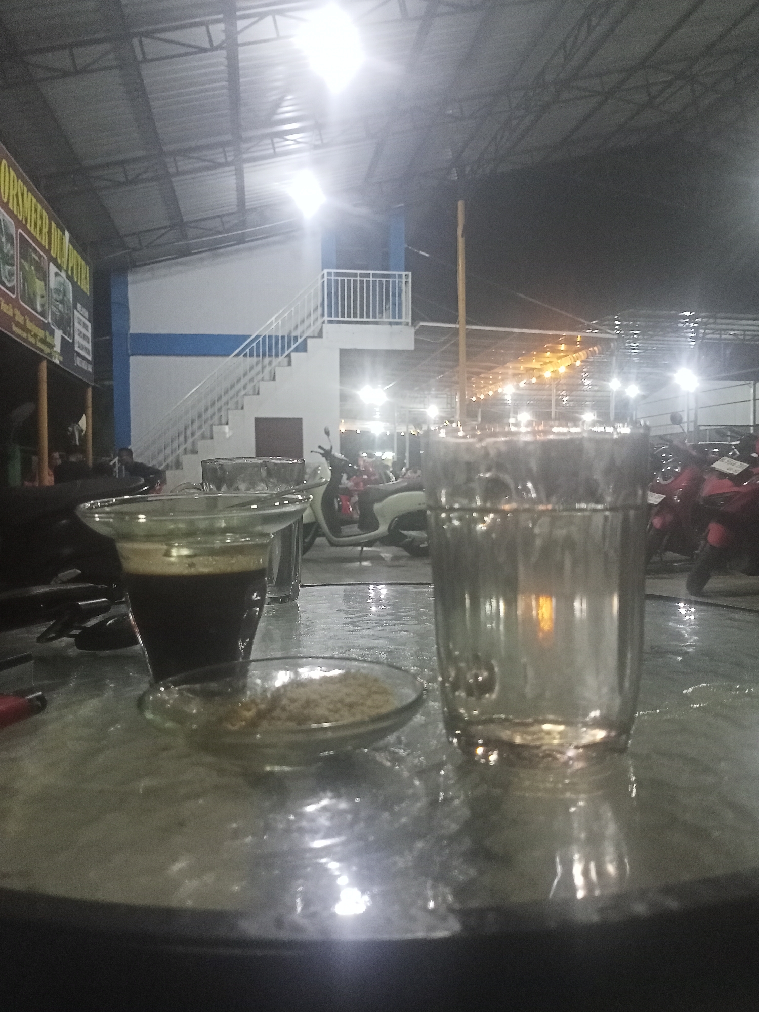 This nighttime image depicts an open-air, covered establishment, possibly a cafe or gathering spot, with bright artificial lighting. In the foreground, a glass table holds two beverages and a small dish. To the left, a glass mug contains a dark liquid, likely coffee, with a layer of foam on top. Next to it, a small, clear glass dish holds a granular, light brown substance, possibly sugar. To the right, a tall, clear glass is filled with water, reflecting the surrounding lights.
In the midground, a building features a prominent white staircase with blue trim, leading to an upper level. The ceiling above is a metal truss structure with a corrugated roof. Several motorcycles are parked in the background and midground, suggesting a frequented area or a place associated with vehicle services. A blurry figure of a person can be seen near the stairs. The entire scene is brightly illuminated by numerous overhead lights, which are visible as strong light sources and reflections, indicating that it is nighttime.
On the left side of the image, a sign is partially visible, bearing the text "ORSMEER DUA PUTRA". The overall atmosphere is casual and well-lit, characteristic of a local hangout or service area.