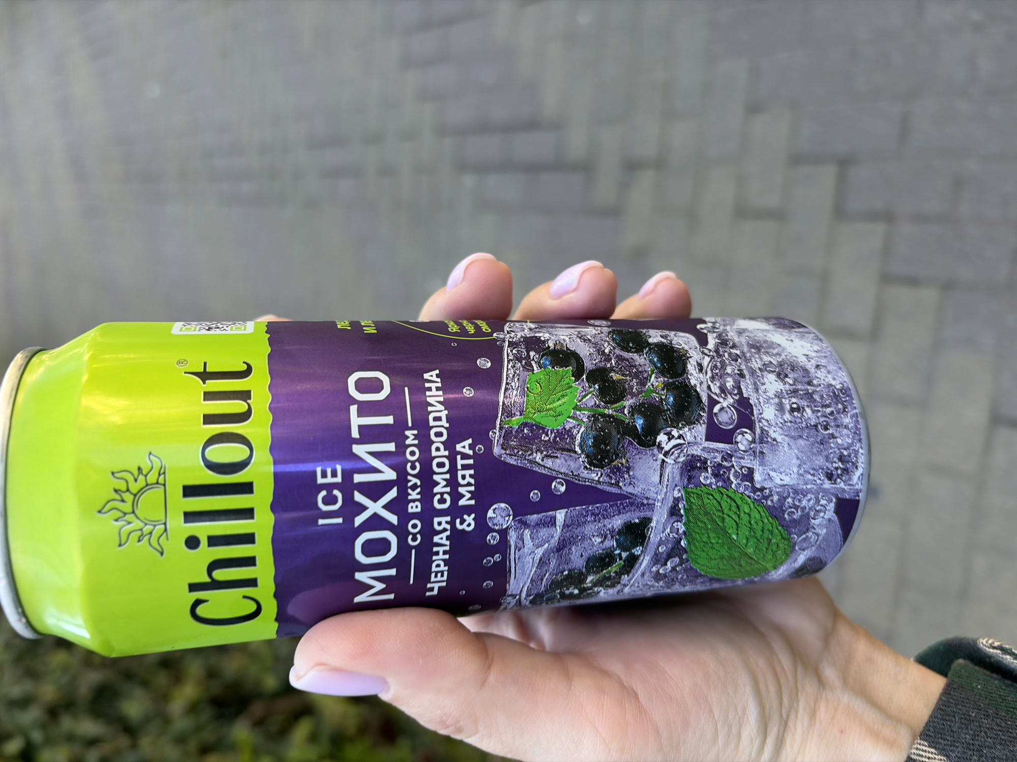 A close-up, eye-level shot captures a person's hand holding a bright green and purple beverage can, likely outdoors in Sochi, Russia. The background is a blurred, grey, paved surface, possibly a wall or ground. The hand shows light pink nail polish on the fingernails.
The can is branded "Chillout" with a sun logo on the green upper section. The lower purple section features the text "ICE МОХИТО" (ICE Mojito). Below this, it specifies the flavor: "СО ВКУСОМ - ЧЕРНАЯ СМОРОДИНА & МЯТА" (WITH THE FLAVOR OF - BLACKCURRANT & MINT). The label design visually represents the flavor with illustrations of ice cubes, blackcurrants, and green mint leaves. Partial text like "ле", "Ил", "C", "Ярко", and "черна" is also visible. The lighting suggests it is daytime.