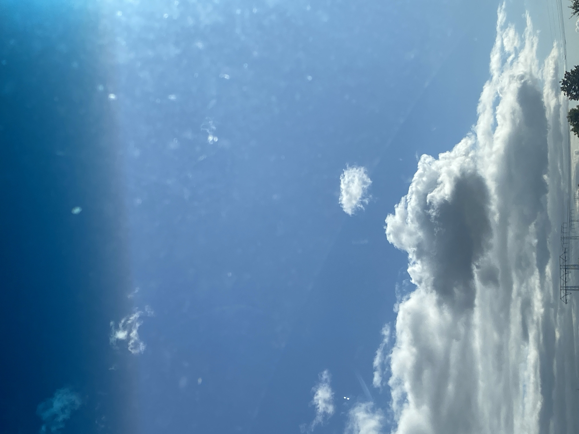 This is an outdoor, eye-level, vertical shot of a partly cloudy sky above Heringen/Helme, Germany. The clouds range from thin, wispy formations to larger, more voluminous cumulus clouds. The sky is a bright blue, with a gradient of lighter blue towards the top of the frame, likely due to the sun's position. There are also some faint, circular reflections or dust particles visible, possibly from the camera lens or a window. In the far distance, there are power lines and the tops of trees, indicating a somewhat rural or suburban landscape beneath the sky. The image captures a moment on a day with dynamic cloud cover and clear skies.