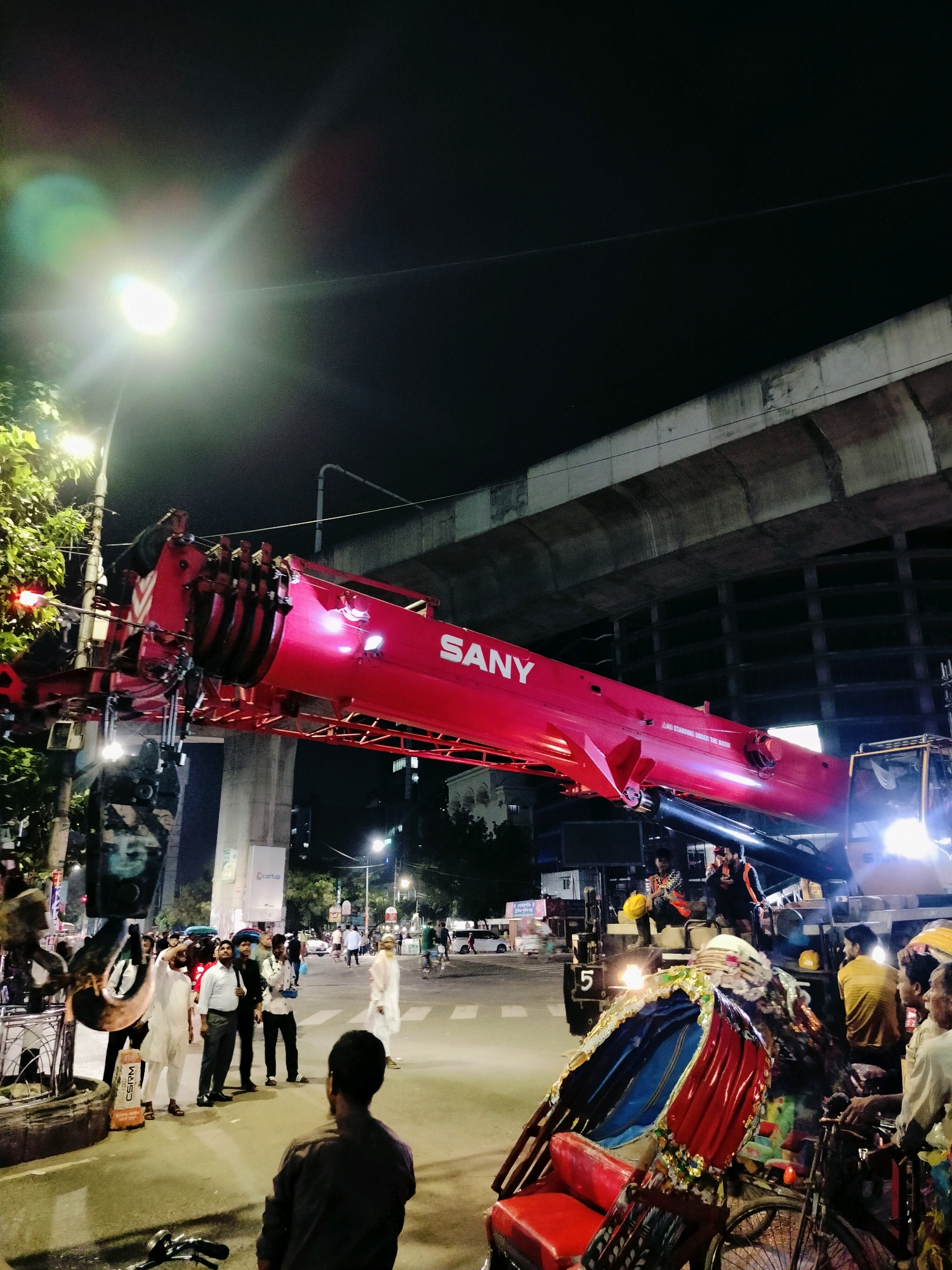 The image depicts a nighttime street scene in Dhaka, Bangladesh, likely near an elevated structure, possibly a flyover or overpass, evidenced by the concrete supports and the dark sky. Dominating the center of the frame is a large, bright red SANY crane, its extended boom stretching across the upper portion of the image. The crane appears to be positioned on a street, with traffic lights and streetlights illuminating the area.
Several people are visible on the street. In the foreground, to the left, a person in a brown shirt is facing away from the camera, appearing to observe the crane. Further back, a group of men in traditional clothing, including white lungis and shirts, are gathered. One man in a white outfit raises his hand. To the right, a colorful rickshaw, a common mode of transport in Dhaka, is parked, with some individuals seated on or around it.
The street is paved and appears to be an intersection, with road markings visible. Other vehicles, including cars and what seems to be a truck associated with the crane, are present but less prominent. The overall atmosphere suggests an active or halted construction or maintenance operation at night, with the powerful crane as the central subject. The lighting conditions, with bright artificial lights and the dark sky, create a dramatic visual effect.