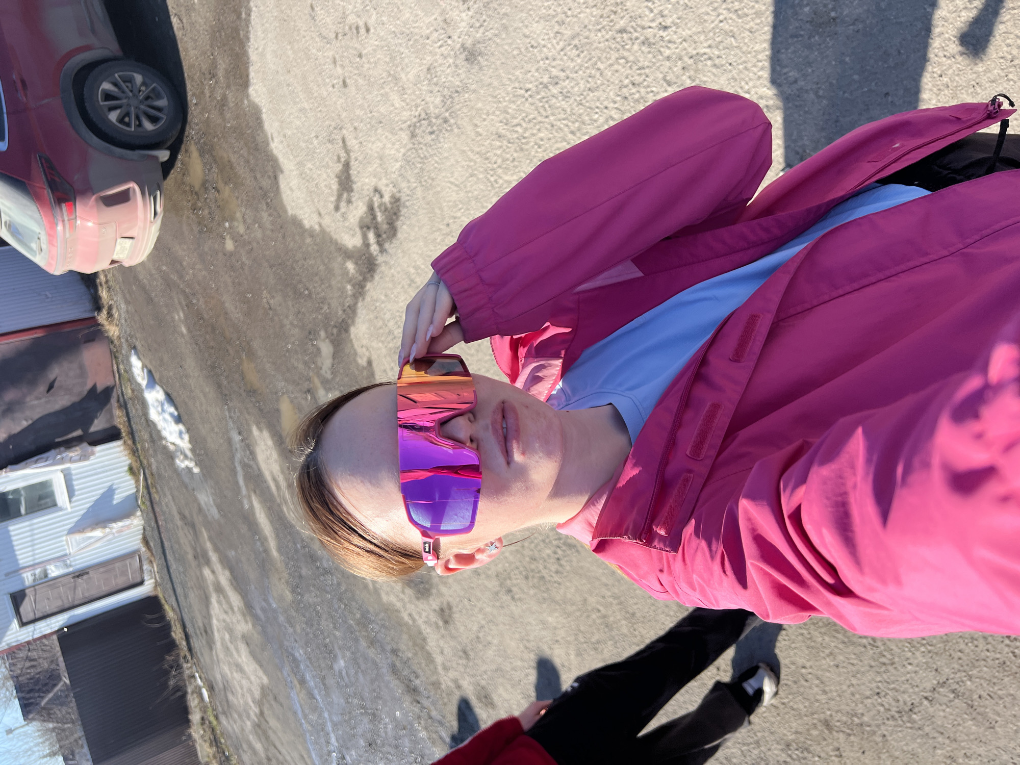 A person is posing for a selfie outdoors on an asphalt surface, likely a street or parking area. The person is wearing mirrored sunglasses with pink and purple lenses, a light blue top, and a bright pink jacket. They are standing on a paved area, which appears to be a road or walkway. To the left is a building with corrugated metal walls and a car parked next to the building. There are patches of snow in the background. The location is presumed to be near Gory, Russia, based on the prompt.