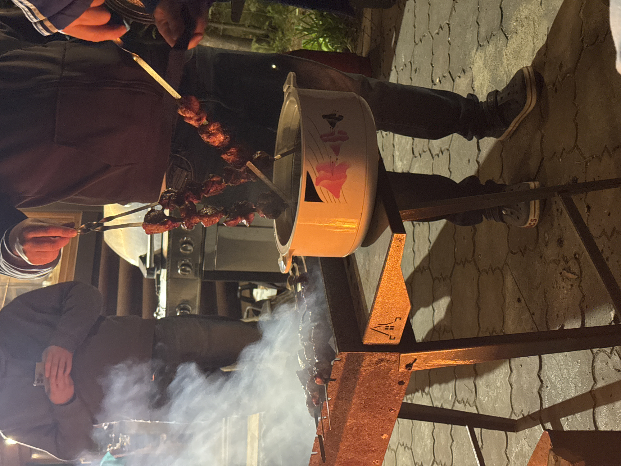 The image shows people grilling meat on skewers in an outdoor setting, likely in the evening. A grill is set up, generating smoke. One person is tending to the skewers. The backdrop shows a paved area, and a building. Given the request, this could be in Tomilino, Russia.