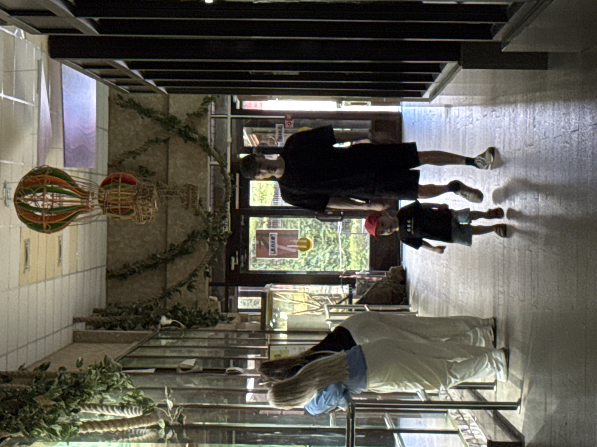 In an indoor walkway, possibly within an exhibition hall or zoo, two adult women with long blonde and brown hair respectively, are looking out through a glass enclosure to their left. They are both wearing light-colored pants, with the blonde woman in a blue top and the brown-haired woman in a dark top. To their right, a man in a black t-shirt, black shorts, and a black baseball cap walks towards the viewer. He is accompanied by a young child wearing a red cap, a dark t-shirt, and light-colored shorts. The child is walking slightly ahead of the man. The walkway features textured flooring and is decorated with artificial greenery hanging from the ceiling and walls. In the background, doors are visible, leading to an outdoor area with greenery. A large, colorful hot air balloon decoration hangs from the ceiling overhead. The location is identified as Izhevsk, Russia.