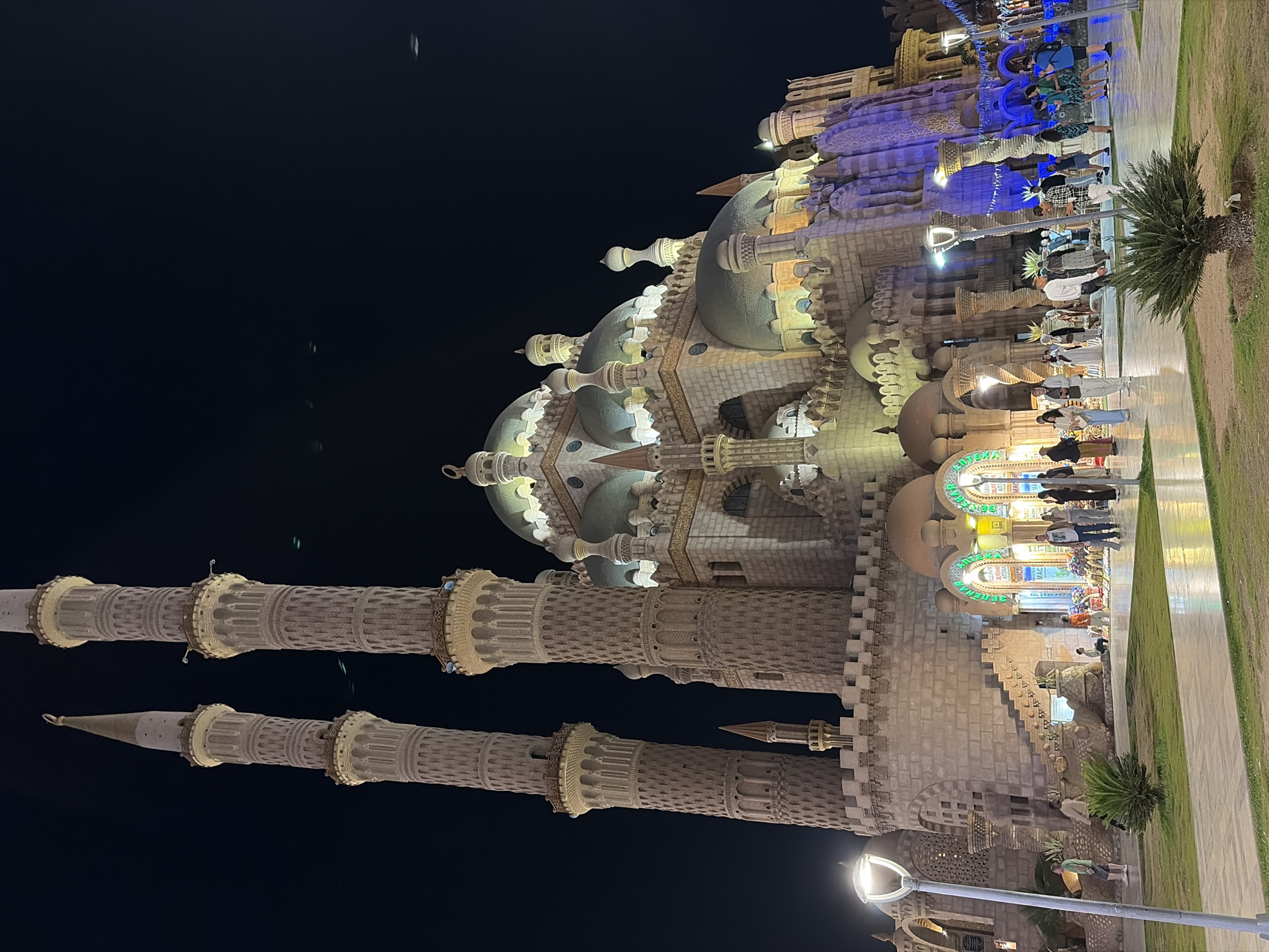 The image depicts the Al Sahaba Mosque in Sharm El Sheikh, Egypt, at night. The mosque is a large, elaborate stone structure illuminated by artificial lights, rendering the sky a deep black. Two tall, ornate minarets with multiple balconies and spires rise prominently on the left, displaying intricate patterns. The main body of the mosque features numerous domes of varying sizes, some with a subtle greenish or bluish tint from the lighting, and arched entrances. The stone facade is a light, sandy color, contrasting with the dark sky.
In the foreground, a wide, light-colored paved plaza extends from the base of the mosque. Patches of green grass with small, palm-like plants are interspersed along the paved areas. Modern streetlights with glowing white luminaires are positioned along the plaza, casting light on the ground and surrounding areas.
Numerous individuals are present on the plaza, walking and standing. They appear to be a mix of adults, dressed in casual attire. On the right side of the mosque's base, several storefronts are visible, brightly lit with arched entrances and displaying some signage in Arabic and Cyrillic script, indicating commercial activity. Merchandise is visible outside some of these shops. People are observed near these shops and strolling across the plaza, some looking towards the mosque, others moving along the paths. The scene is active, suggesting a public space frequented at night.