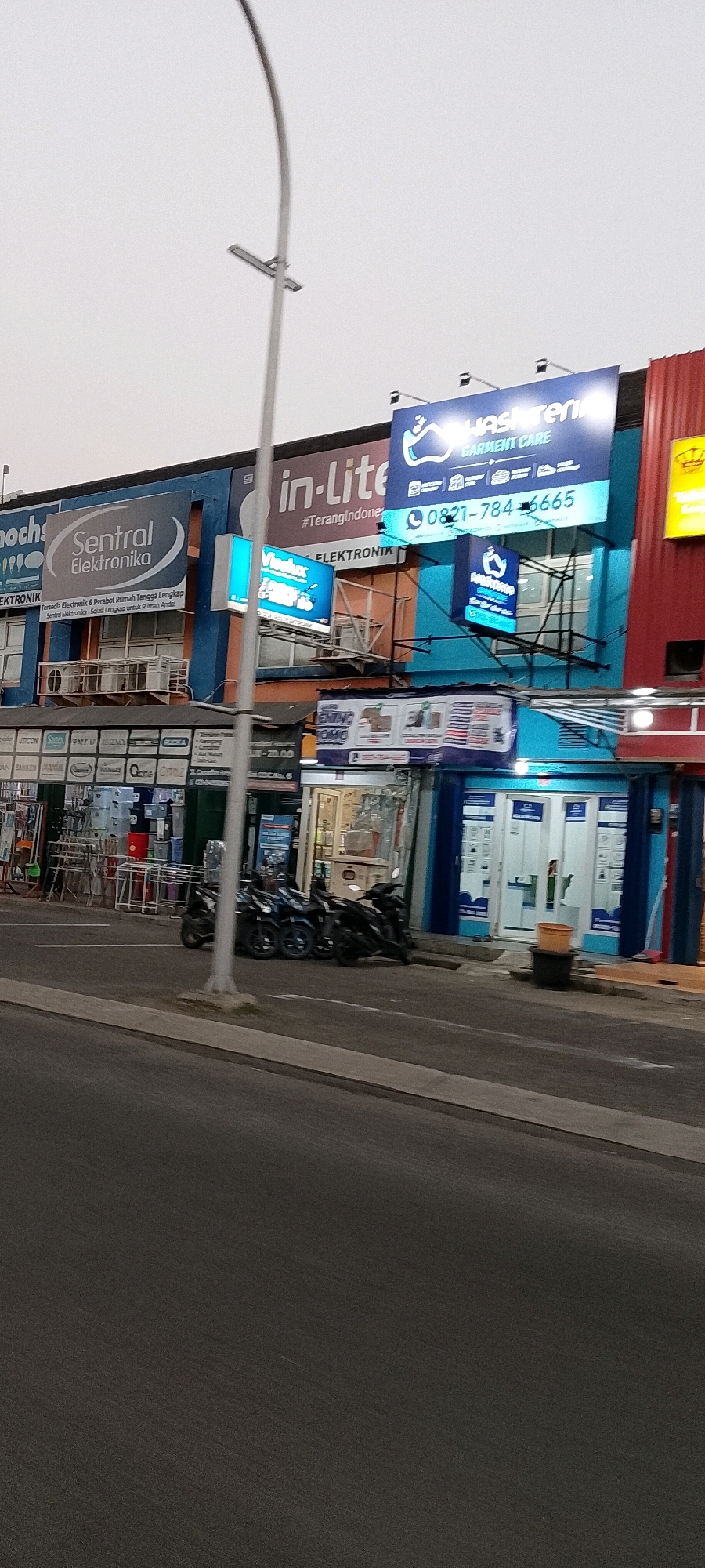 A commercial building facade in Cileungsi, Indonesia, is captured at ground level, showing multiple storefronts. A prominent sign on the right reads "washeroni GARMENT CARE" with a phone number "0821-784-6665". To the left, a blue sign displays "Sentral Elektronika" with smaller text below. Another sign, also blue, reads "in-Lite #TerangIndonesia". The building features windows and signage for various businesses. Several motorcycles are parked on the sidewalk in front of the shops. The street in the foreground is paved and a sidewalk runs along the building. The sky is overcast.