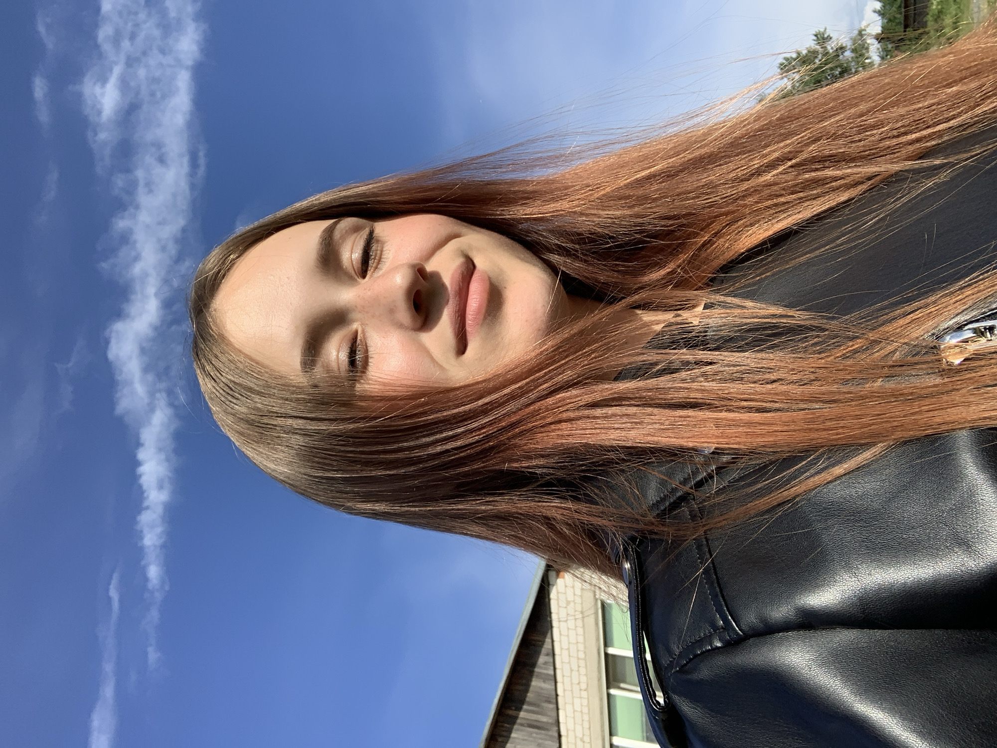 A young woman is captured in a close-up, slightly upward-facing selfie, rotated 90 degrees counter-clockwise. She has long, reddish-brown hair that appears to be gently blowing, and is wearing a black leather jacket. Her eyes are mostly closed, and a subtle, content smile plays on her lips, suggesting she is enjoying the bright sunlight. Freckles are visible across her nose.
The background is dominated by a clear, vibrant blue sky with wispy white clouds trailing across it. In the bottom right corner (when the image is oriented correctly), part of a building with light-colored brick and dark wood siding, along with some windows, is visible. Green foliage from distant trees can be seen in the upper right. The lighting is bright and natural, indicating a sunny daytime setting. The overall mood is peaceful and happy, reflecting enjoyment of the outdoor environment. There is no visible text in the image. The scene takes place in Фоки, Russia.