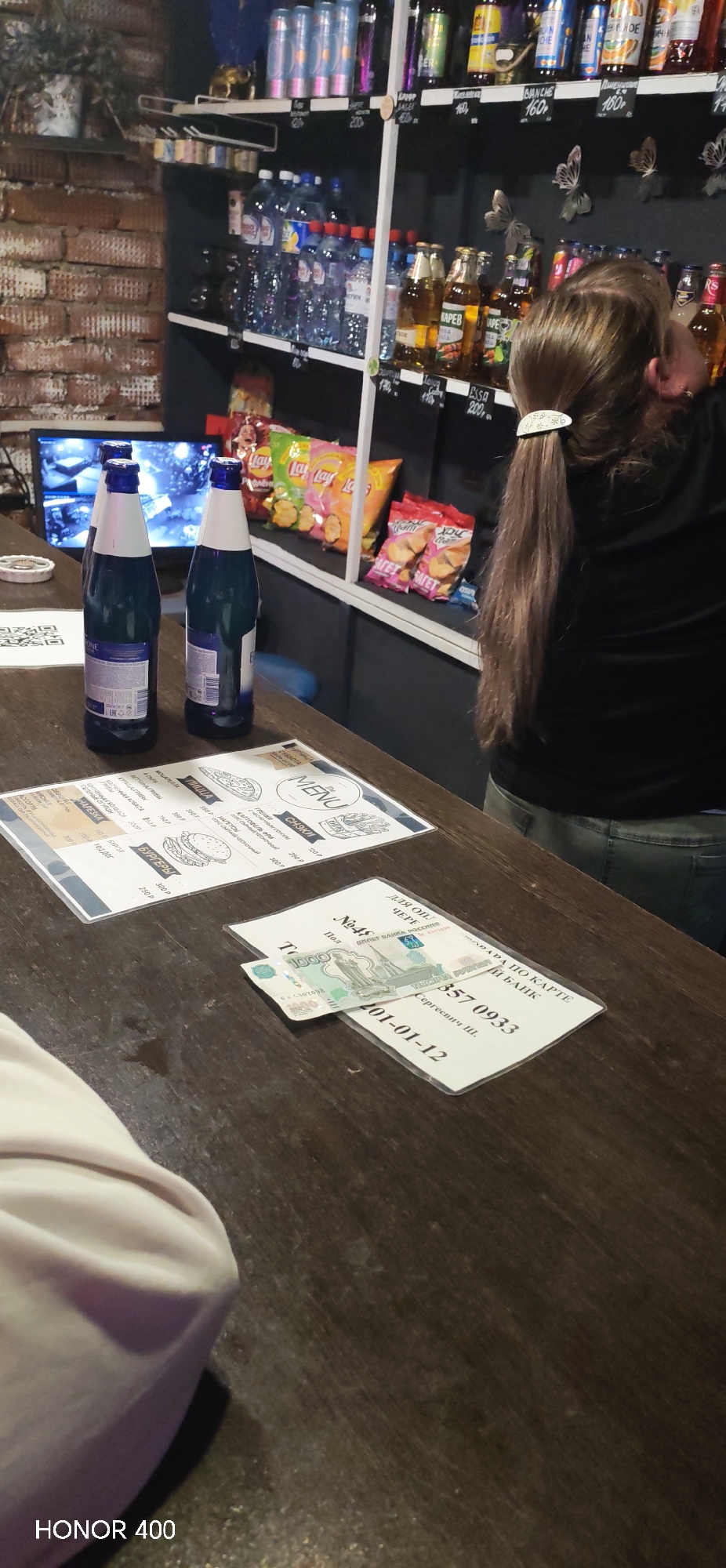 A close-up shot inside a bar in Yakshur-Bod'ya, Russia, presents a scene on a wooden countertop. Two blue glass bottles stand behind a menu card and a payment receipt, on top of which lies a 1000 Ruble bill. A woman stands behind the counter, partially obscured, wearing a black top and jeans, with her brown hair pulled back in a ponytail. Shelves behind the counter display various drinks and snacks, including water bottles, beer, chips, and other beverages, with prices indicated on small signs. A television screen is visible in the background, behind the bottles. The environment is illuminated by the bar's lighting.