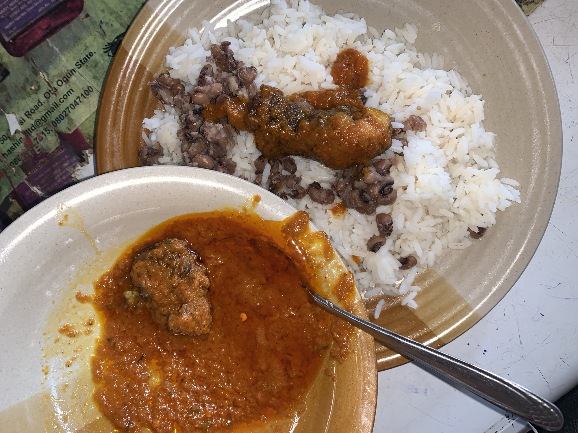 The image shows two plates of food. The plate on the right contains white rice, black-eyed peas, and a portion of meat covered in a brown sauce. The plate on the left contains a portion of meat covered in a red sauce, with a spoon visible next to it. A flyer or document with partial text is visible in the top left, including the address "50, si Road, Ota Ogun State, Nigeria" and a Gmail address, which indicates the photo was possibly taken near Kabong, Nigeria, as it is in Nigeria. The table has a textured, possibly dirty, surface.